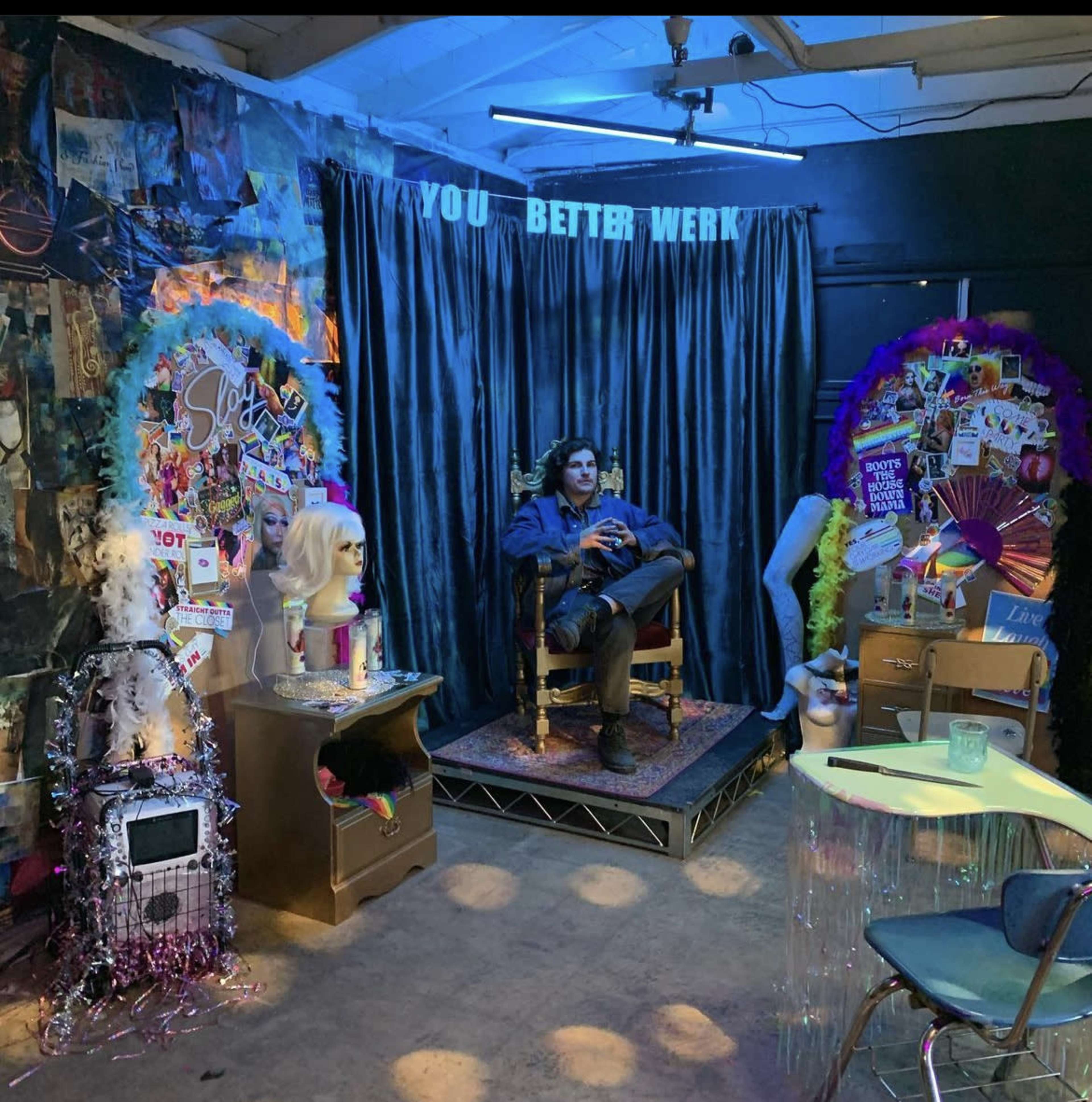 A man sits in a chair on stage surrounded by colorful decorations, with a backdrop that reads "YOU BETTER WORK."