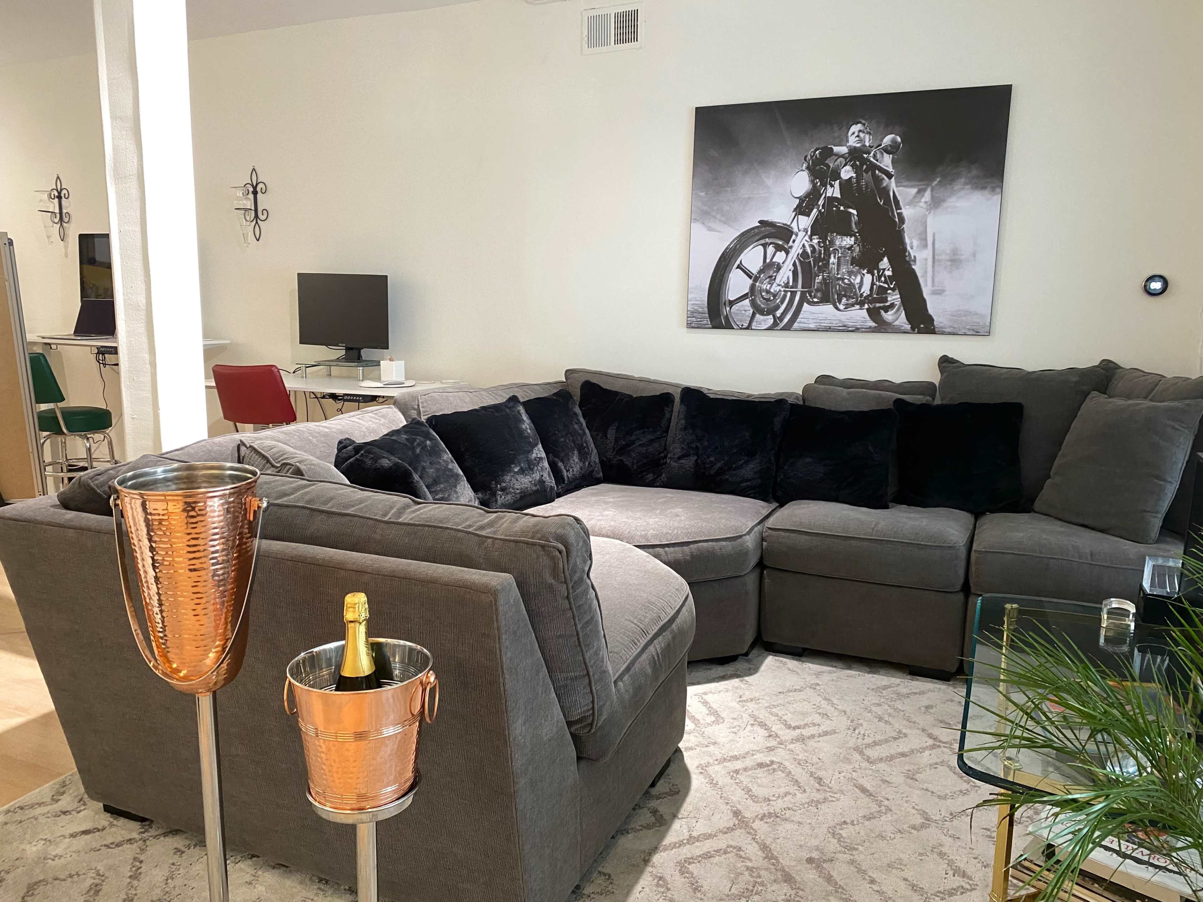 The image shows a modern living room featuring a gray sectional sofa, a decorative wall art of a motorcycle-themed scene, and a beverage cart with a champagne bottle.