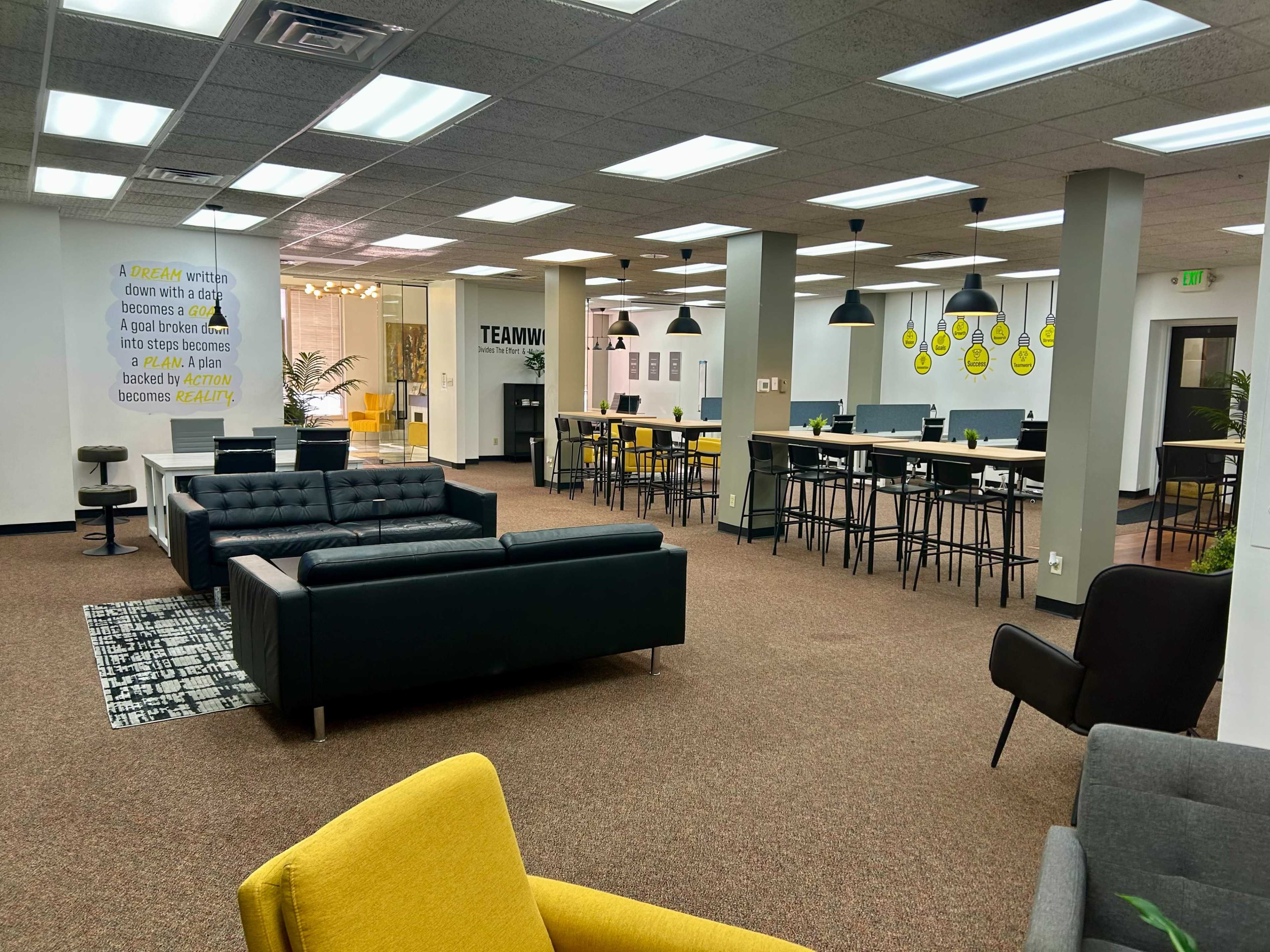 The image shows a spacious, modern office area featuring various seating arrangements, tall tables, and motivational wall art.