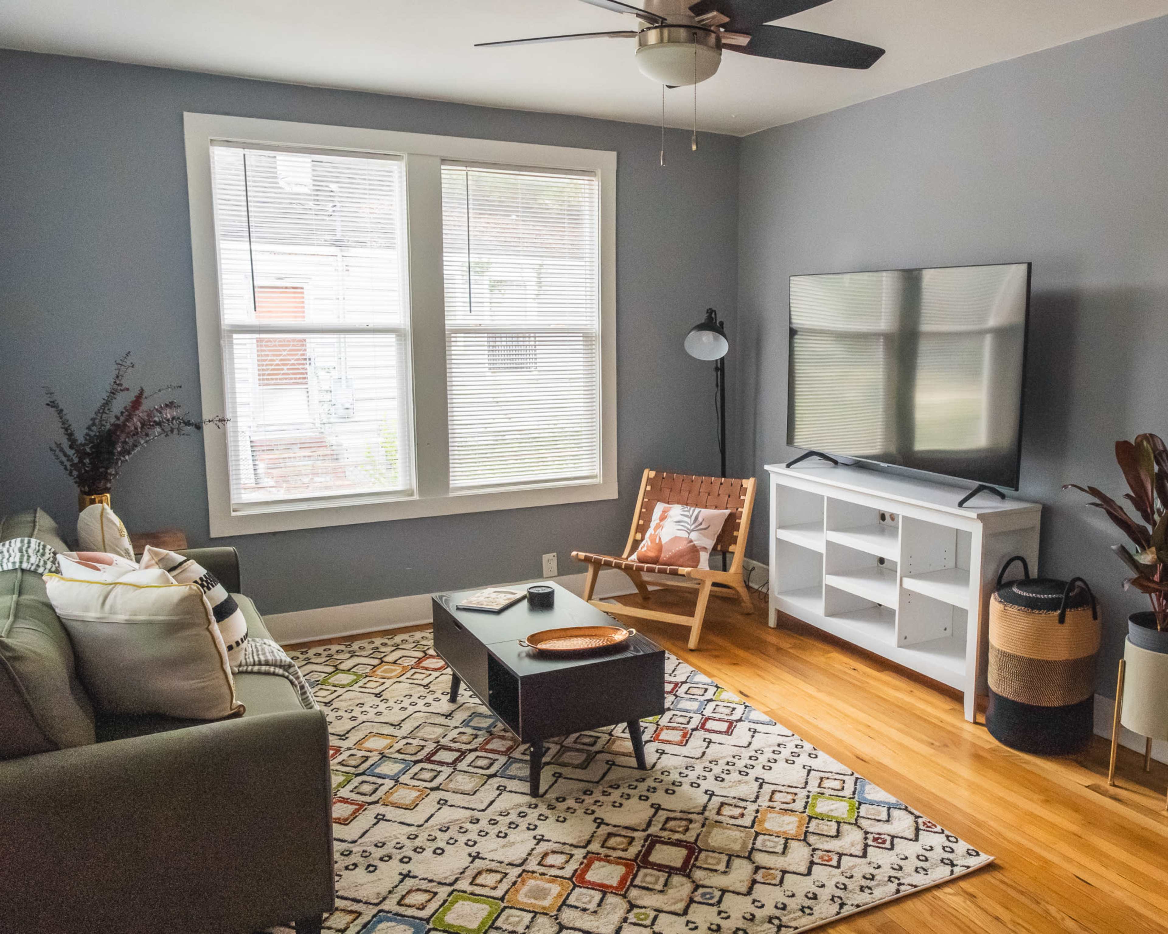 A cozy living room features a green sofa, a coffee table, a television on a white stand, and large windows with natural light.