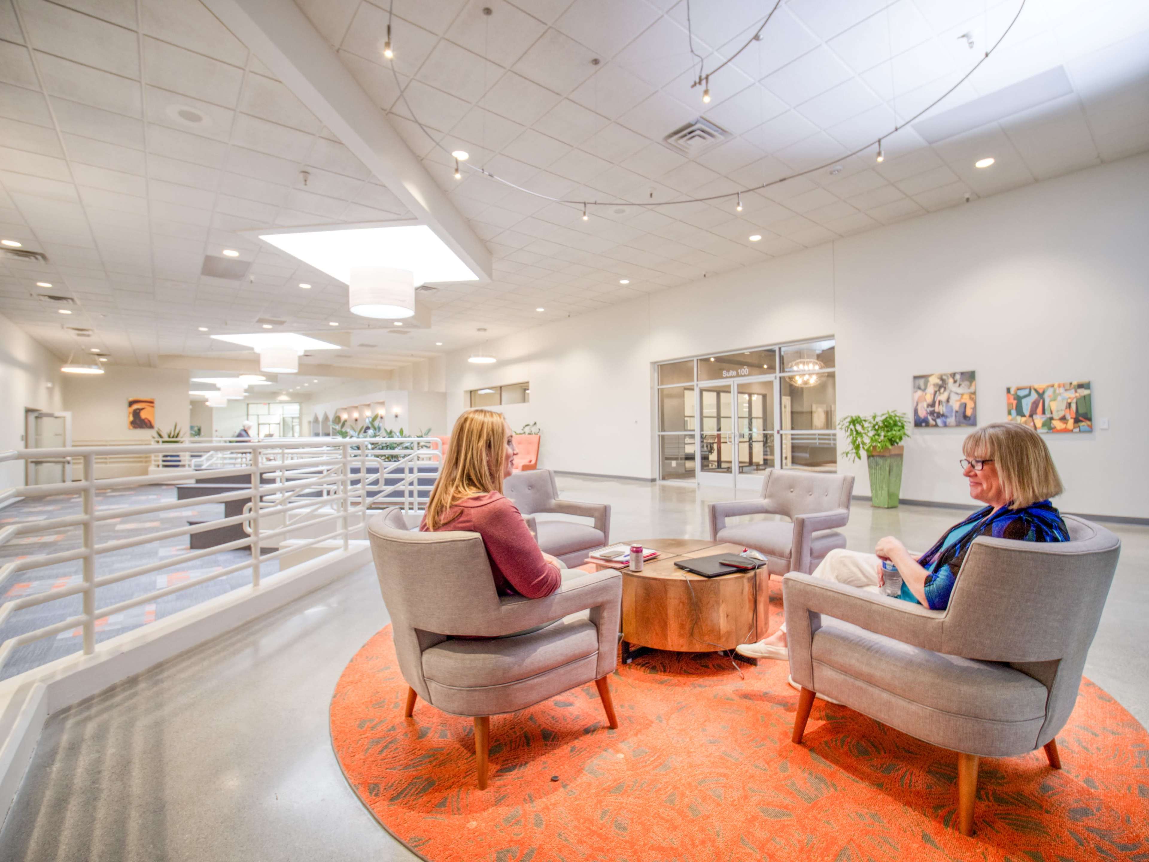 Two women sit in gray chairs around a wooden table on a circular orange rug in a brightly lit modern interior space.