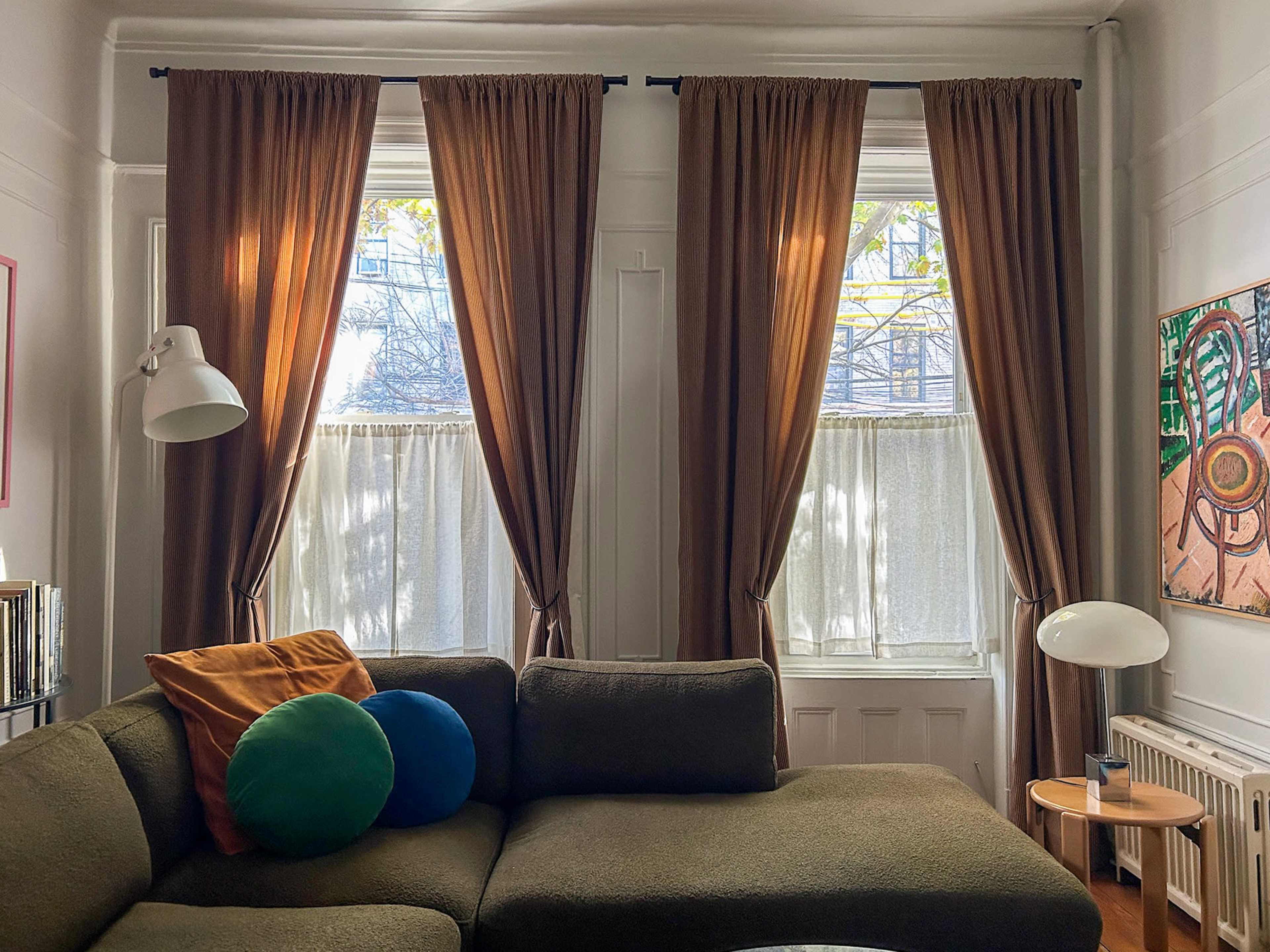 A cozy living room features a large green couch with colorful pillows, flanked by tall windows covered with brown curtains.