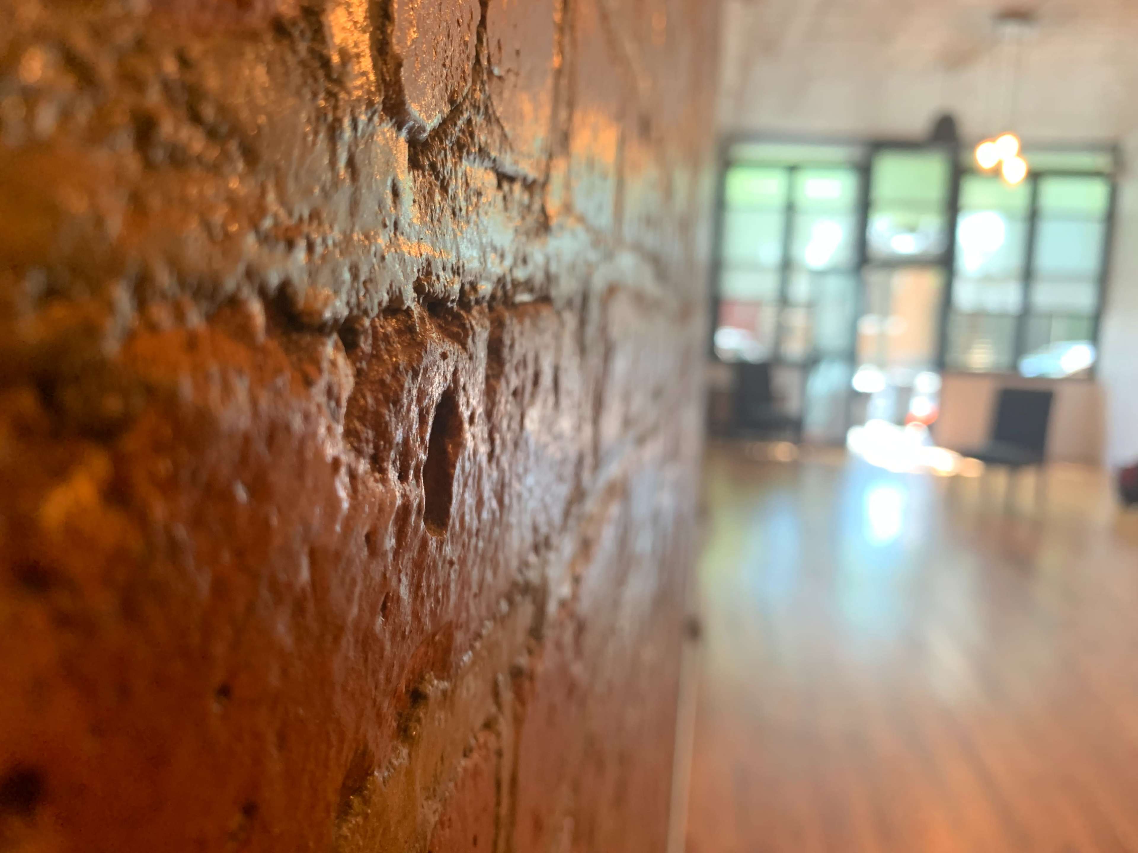 A close-up of a textured brick wall with a blurred view of a well-lit room and large windows in the background.