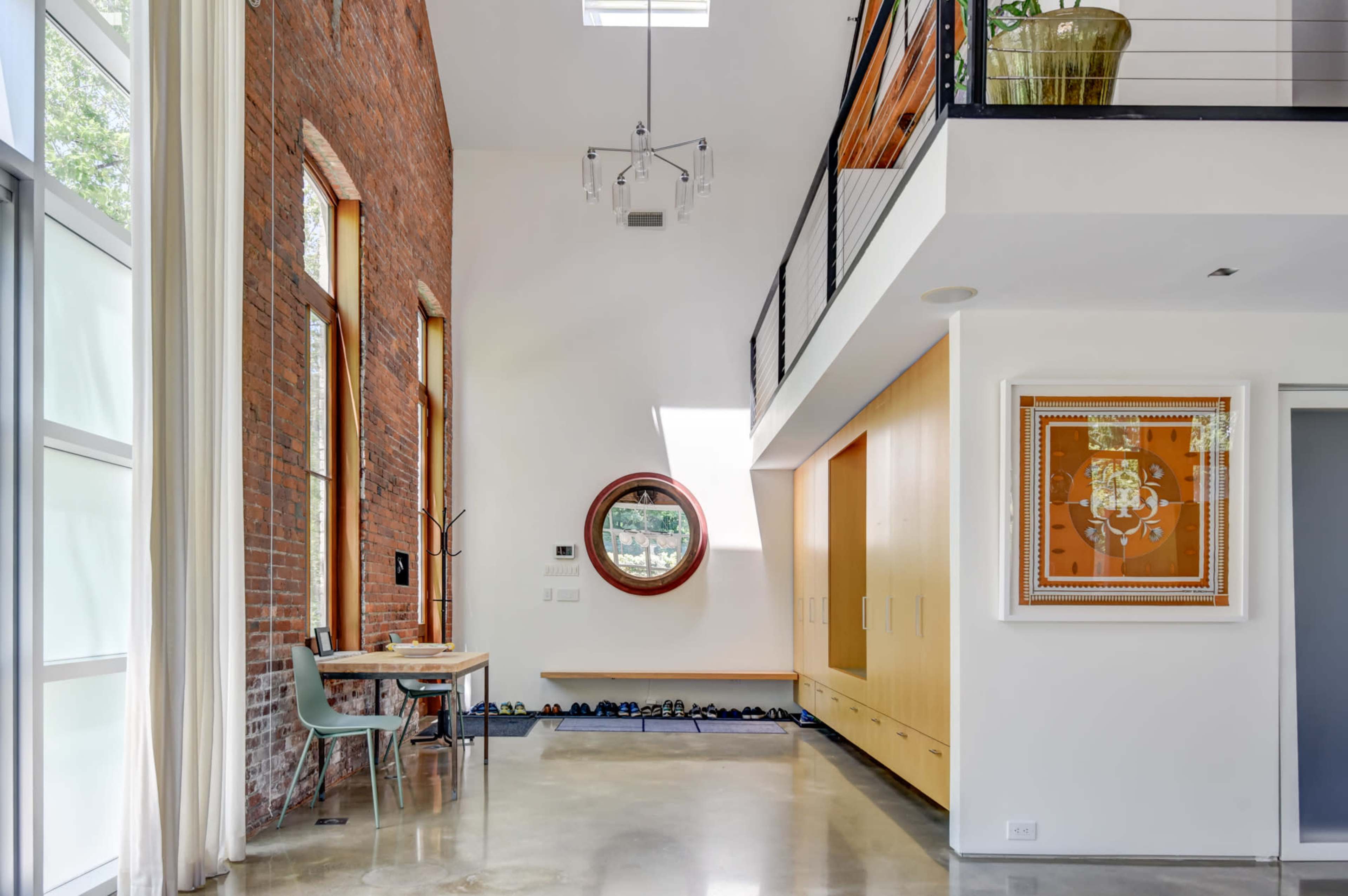 A modern interior space with a high ceiling, featuring a mix of exposed brick and white walls, a circular window, and a wooden table next to a chair.