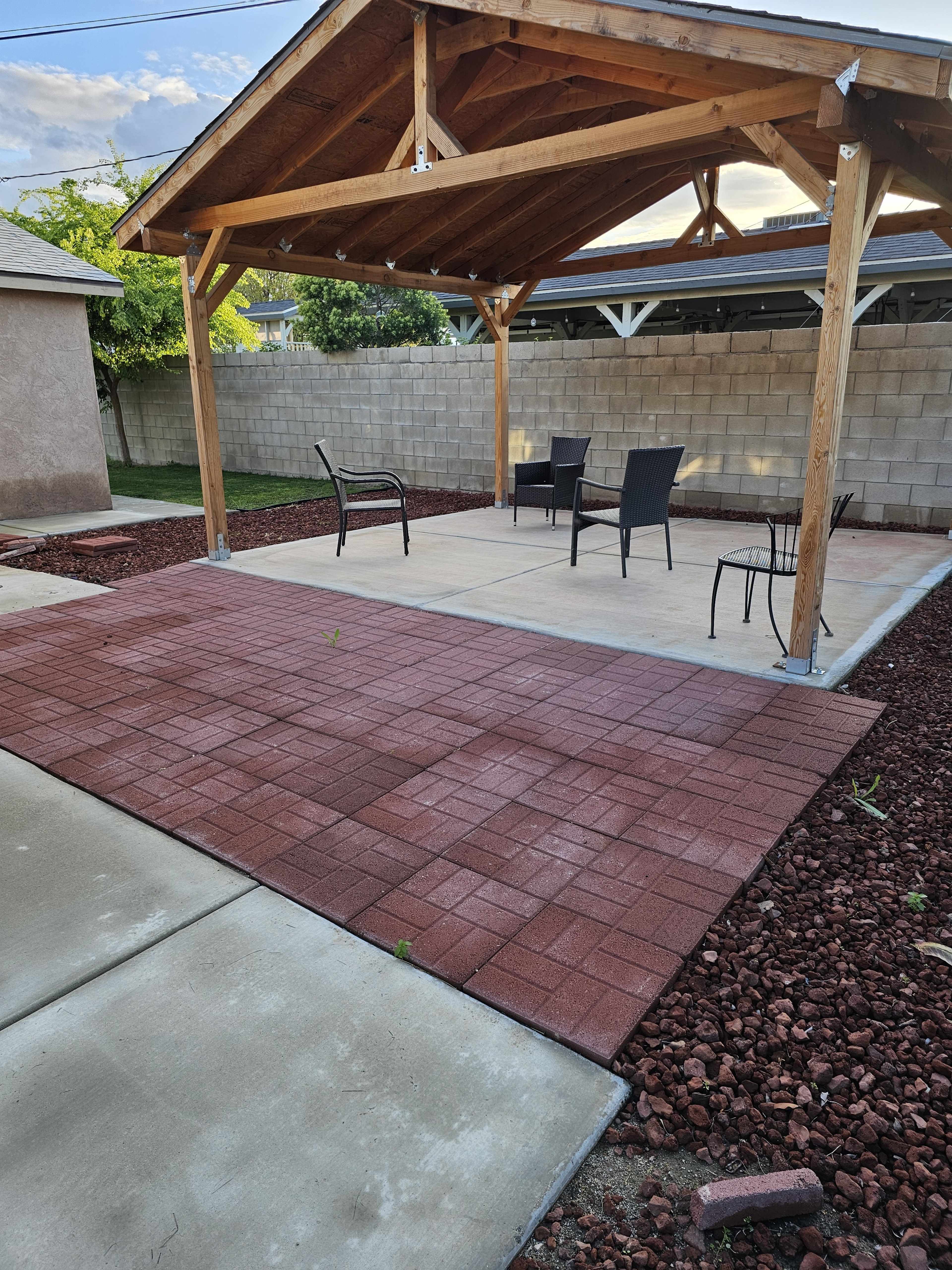 A patio area with a wooden pavilion, surrounded by gravel and featuring several chairs on a concrete surface.