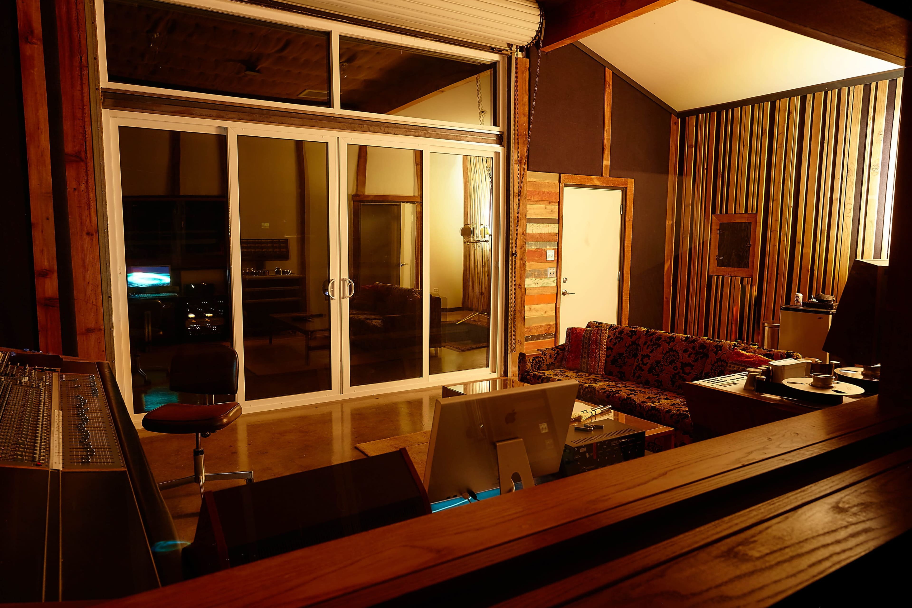 A modern recording studio with a mixing console, a computer workstation, a colorful couch, and large glass doors leading to an outdoor area.