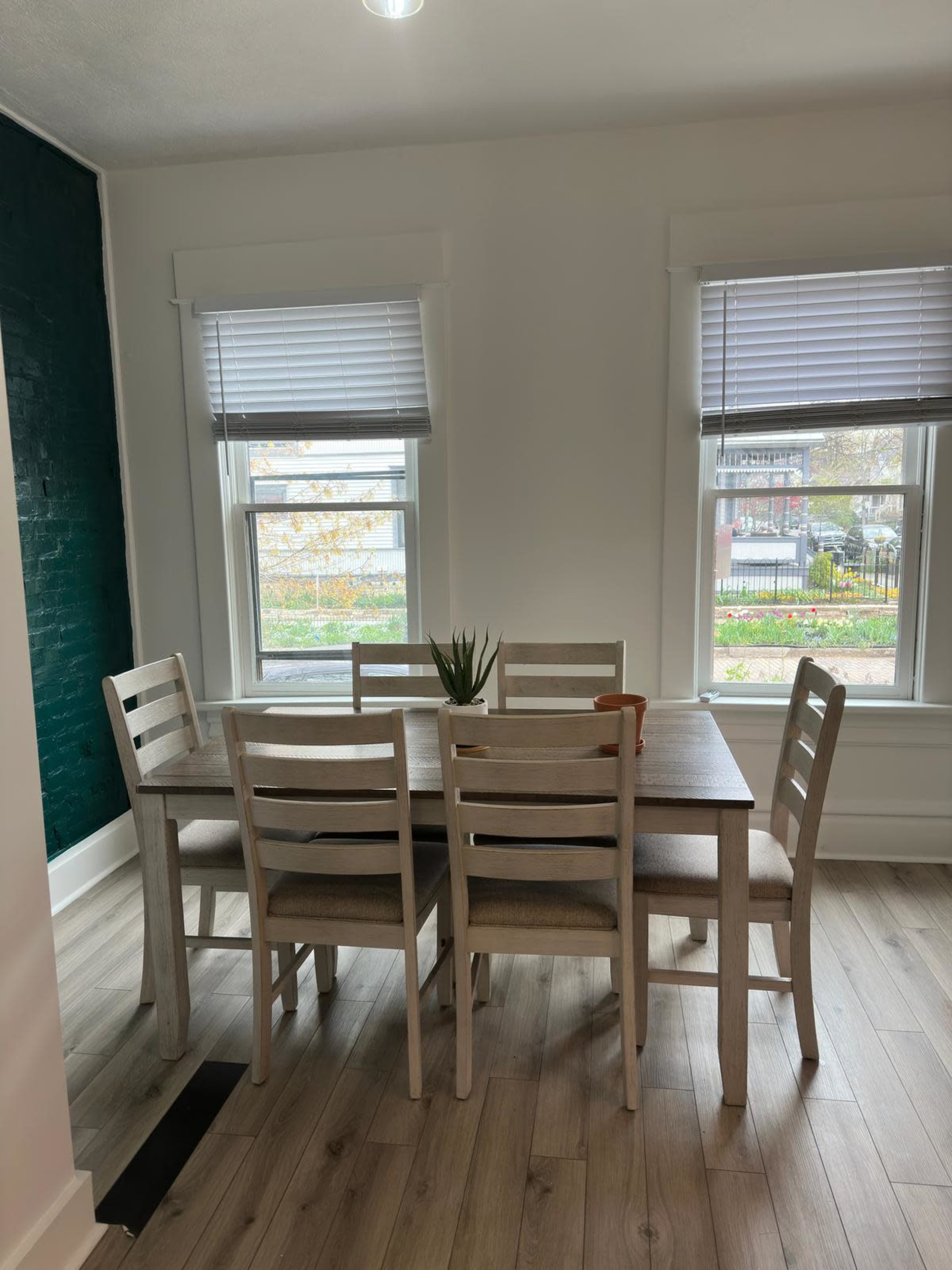 A wooden dining table with six chairs is set in a well-lit room featuring two windows with white blinds.