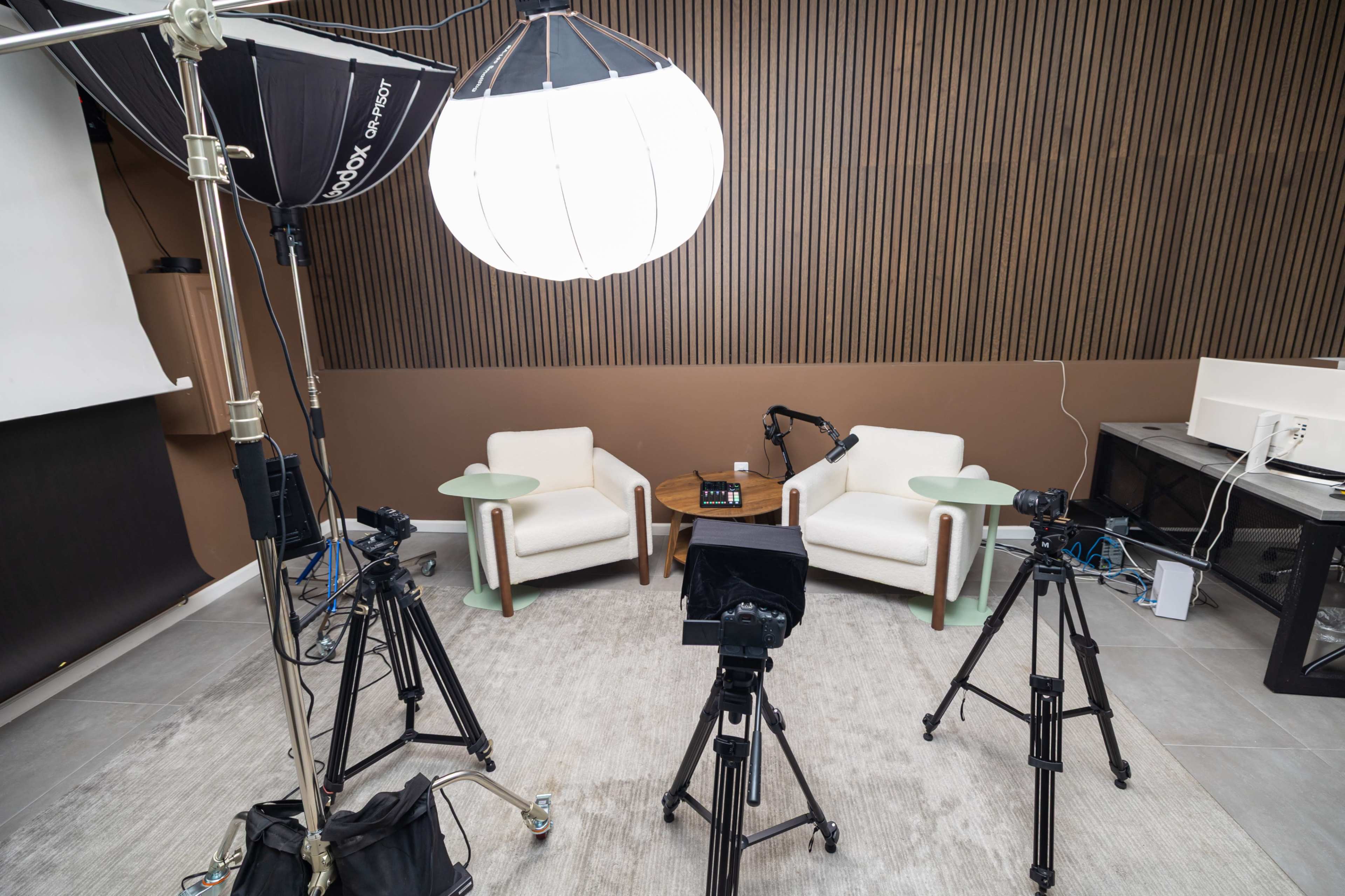 The image shows a studio setup with two white chairs and a small table in the center, surrounded by several cameras, lighting equipment, and a backdrop.