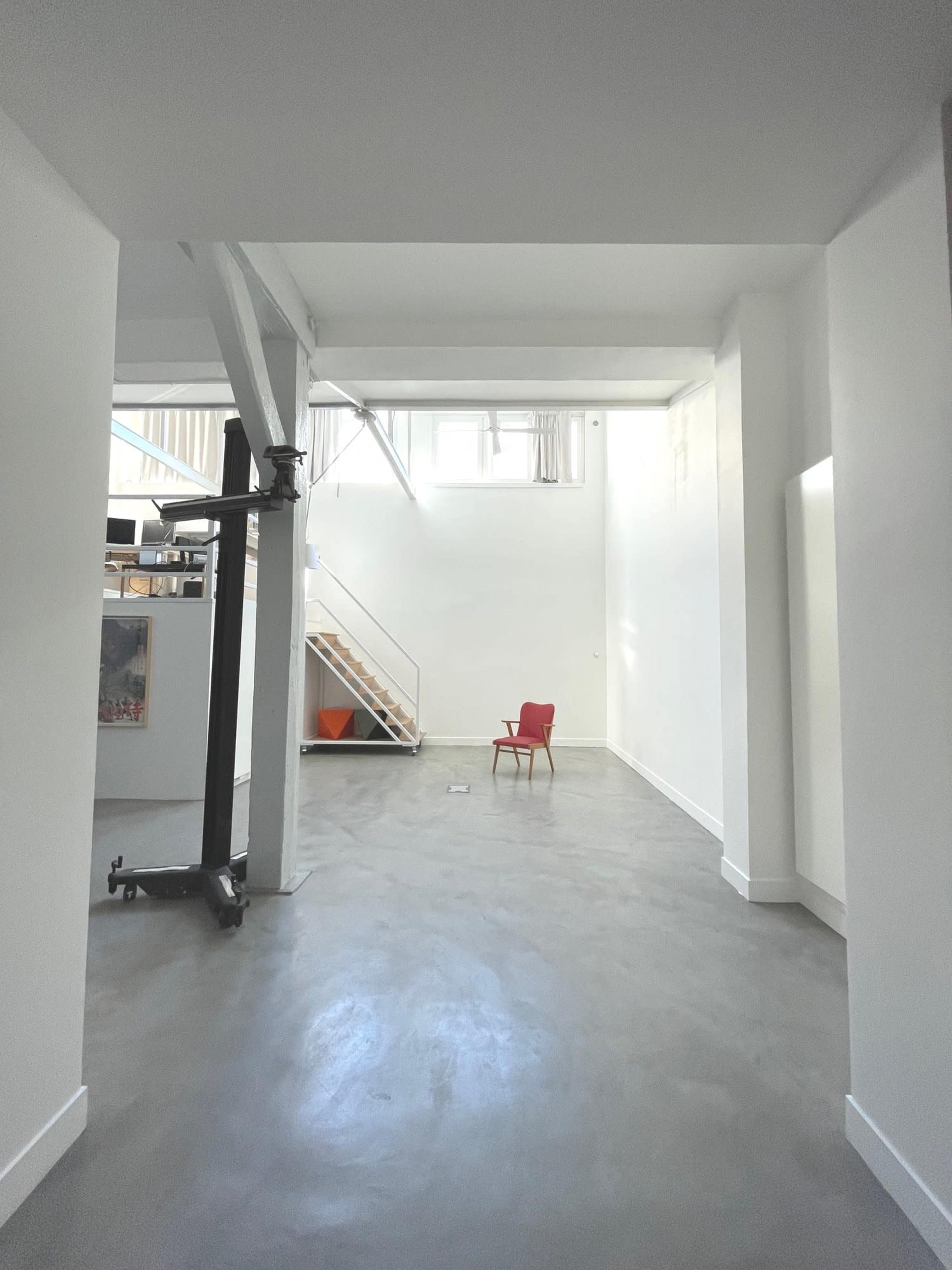 A spacious, minimalist interior with high white walls, exposed beams, and a single red chair positioned in the center.