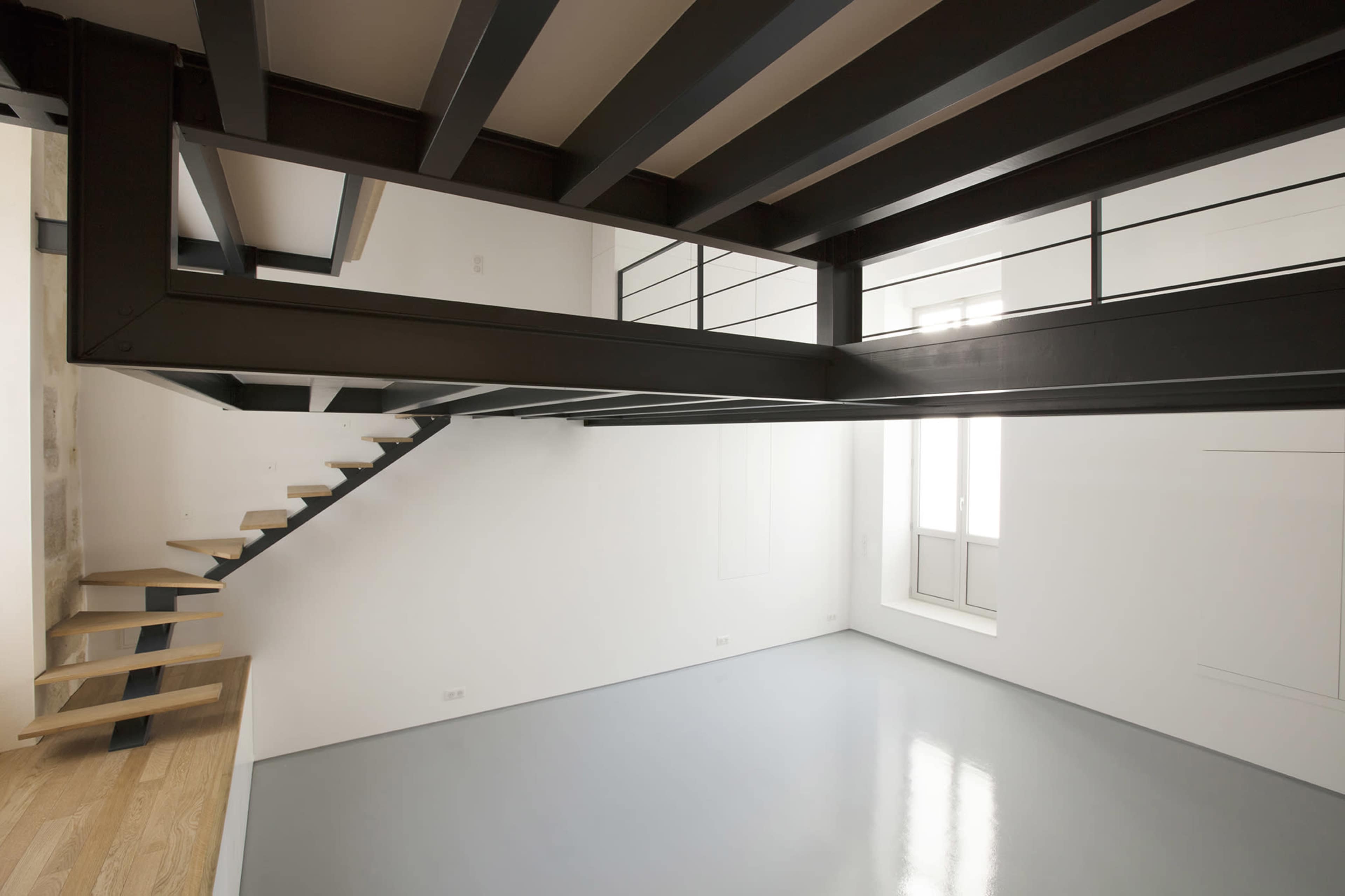 The image shows a modern interior space with an open layout, featuring a steel staircase and a large window allowing natural light to fill the room.