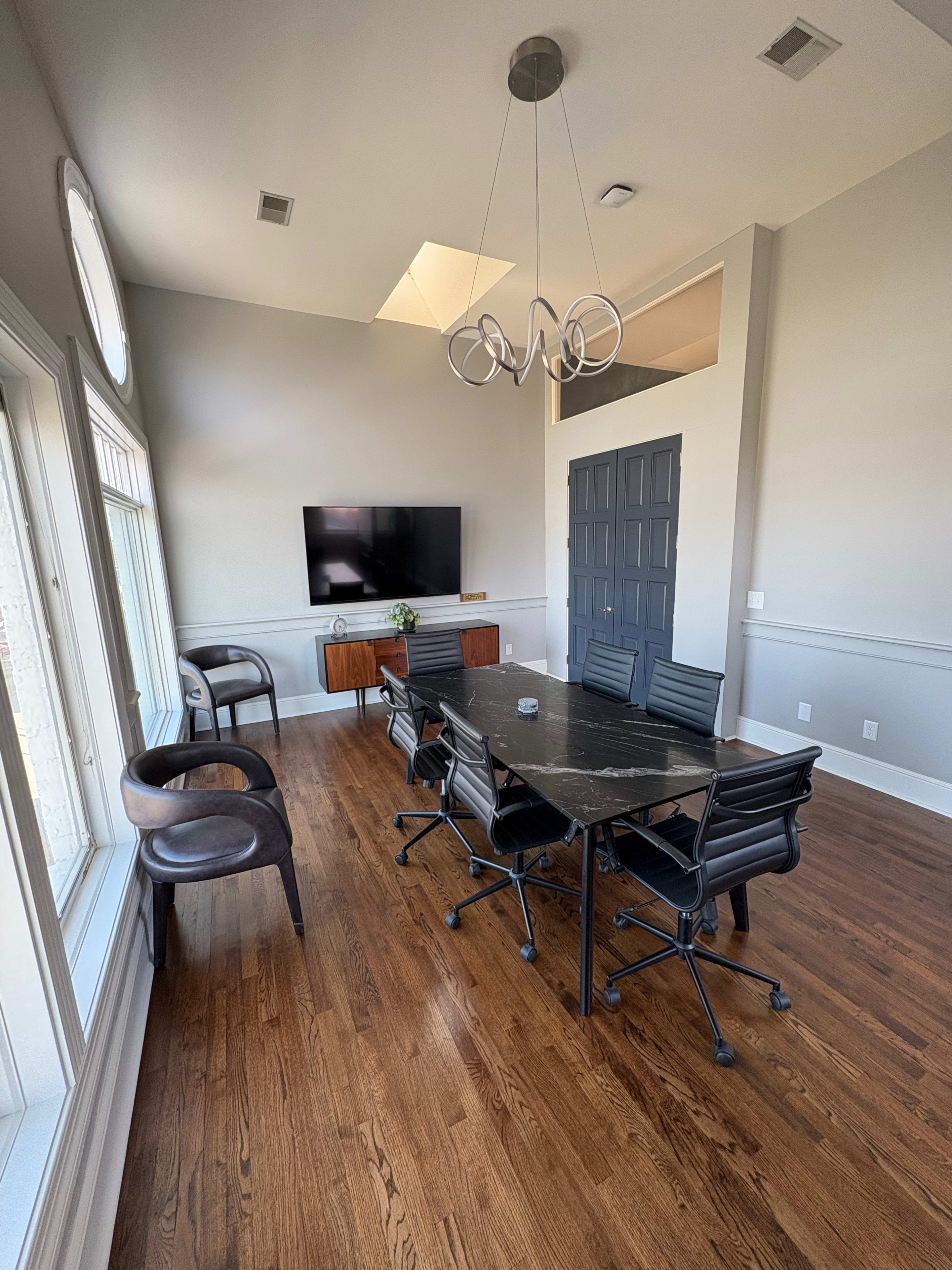 A modern conference room features a large black table surrounded by leather executive chairs, with a wall-mounted television and minimalistic decor.