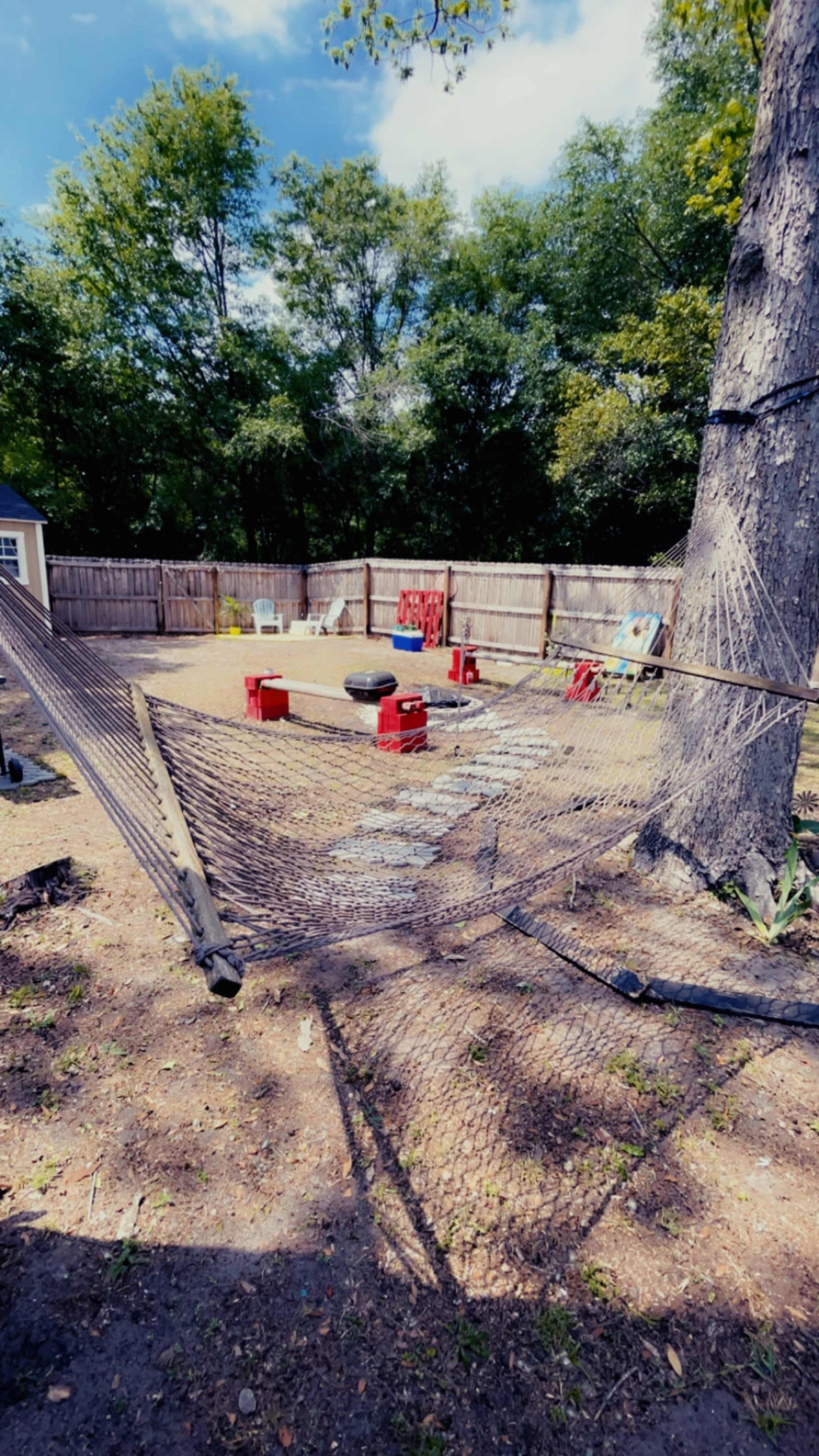 A backyard with a hammock stretched between a tree and a wooden fence, surrounded by red stools and various outdoor items.