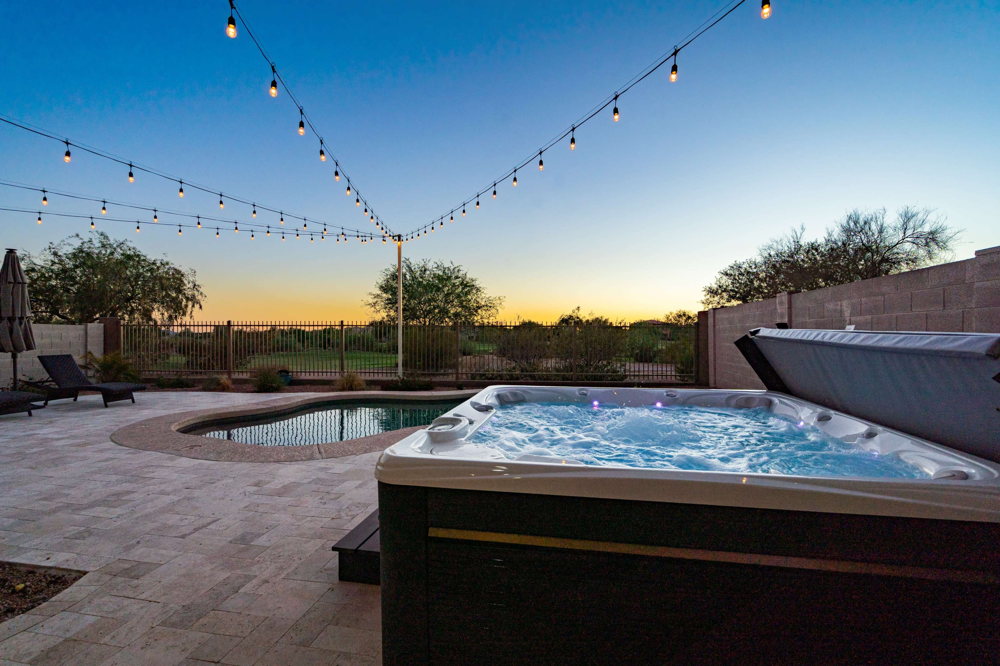 A hot tub filled with bubbling water is situated beside a swimming pool, under string lights against a sunset sky.