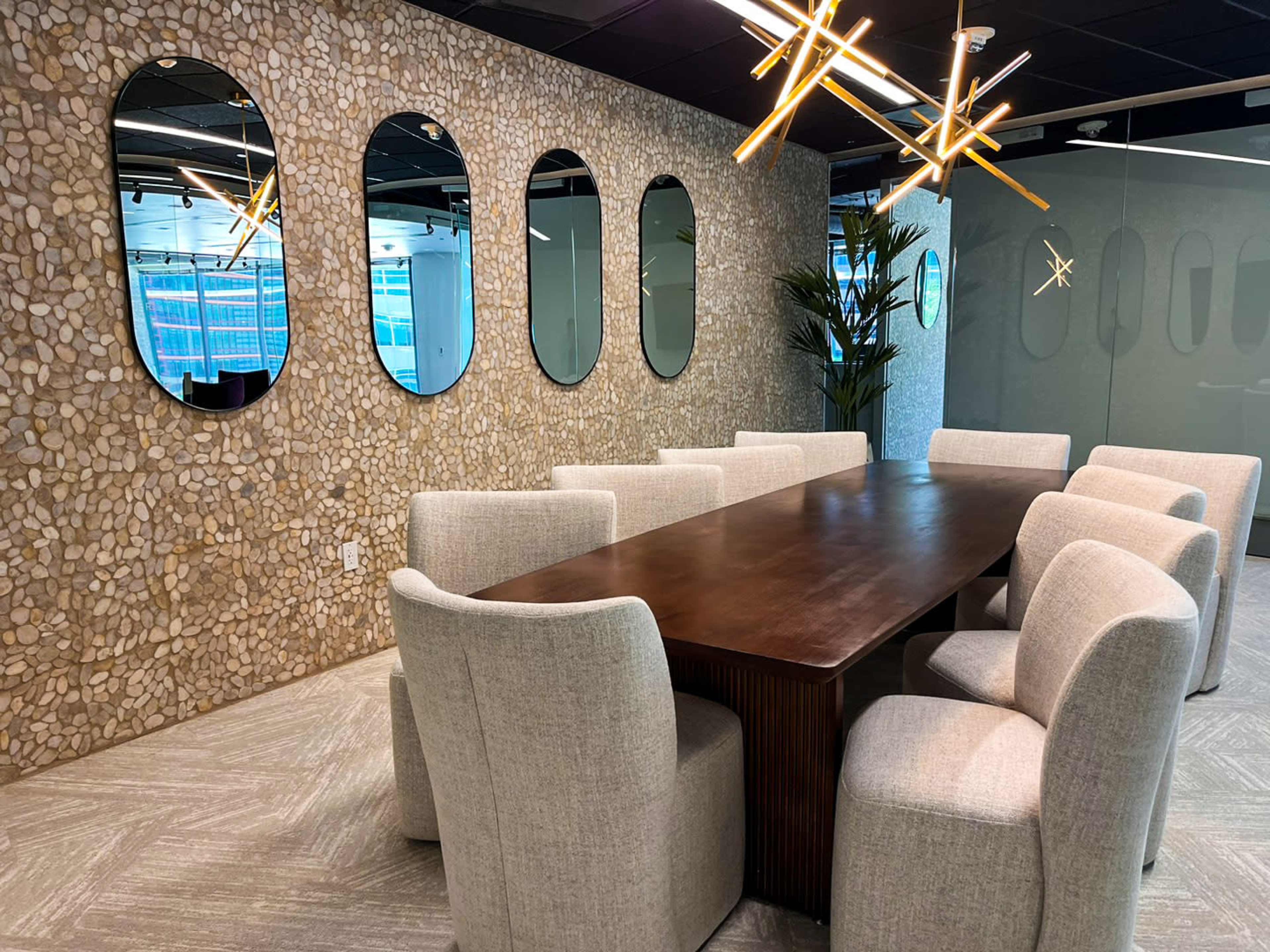 A long wooden table is surrounded by upholstered chairs in a modern conference room featuring a stone accent wall and decorative mirrors.