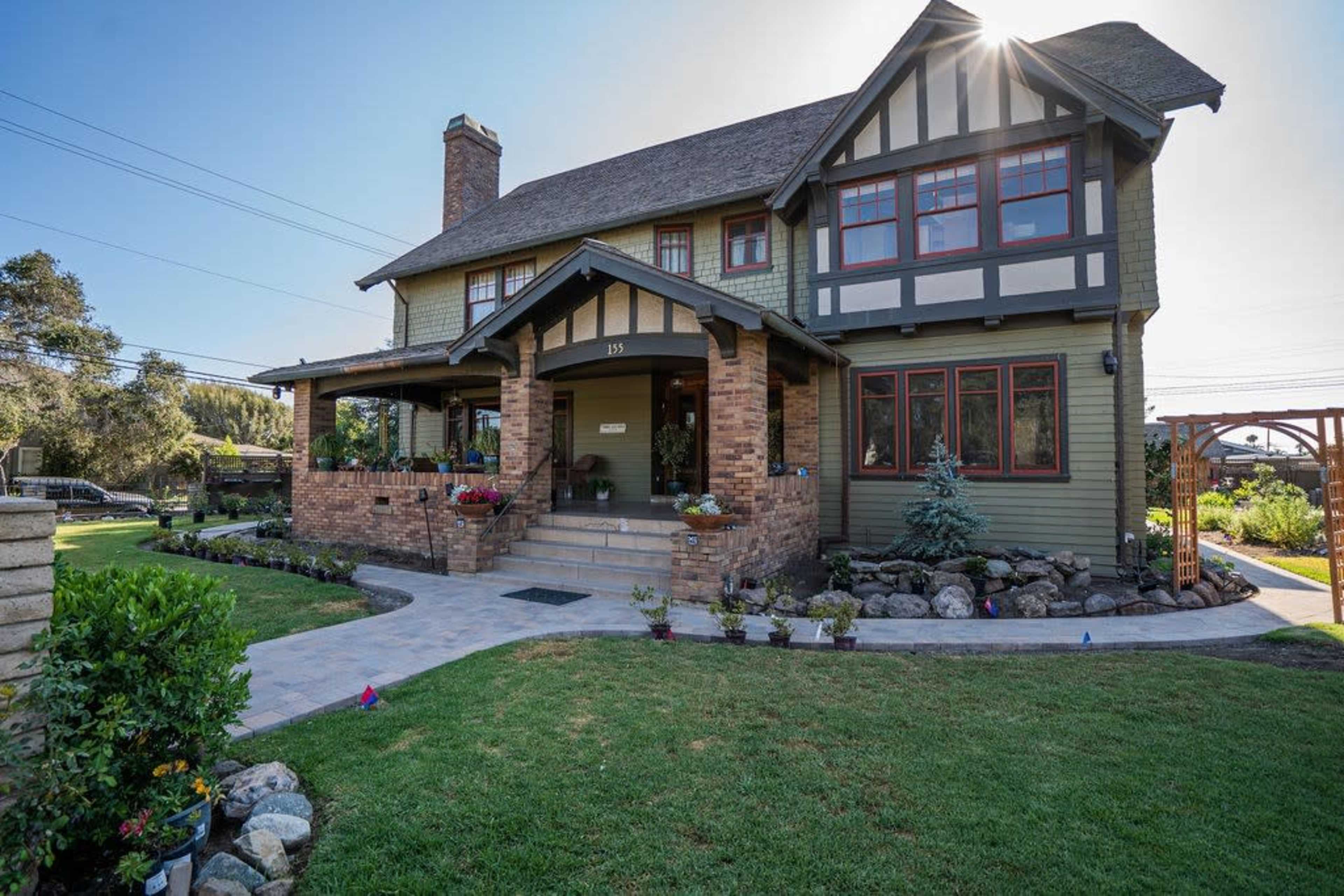 The image shows a large, two-story house with a mix of brick and wood siding, featuring a front porch and a landscaped yard with flowers and small rocks.