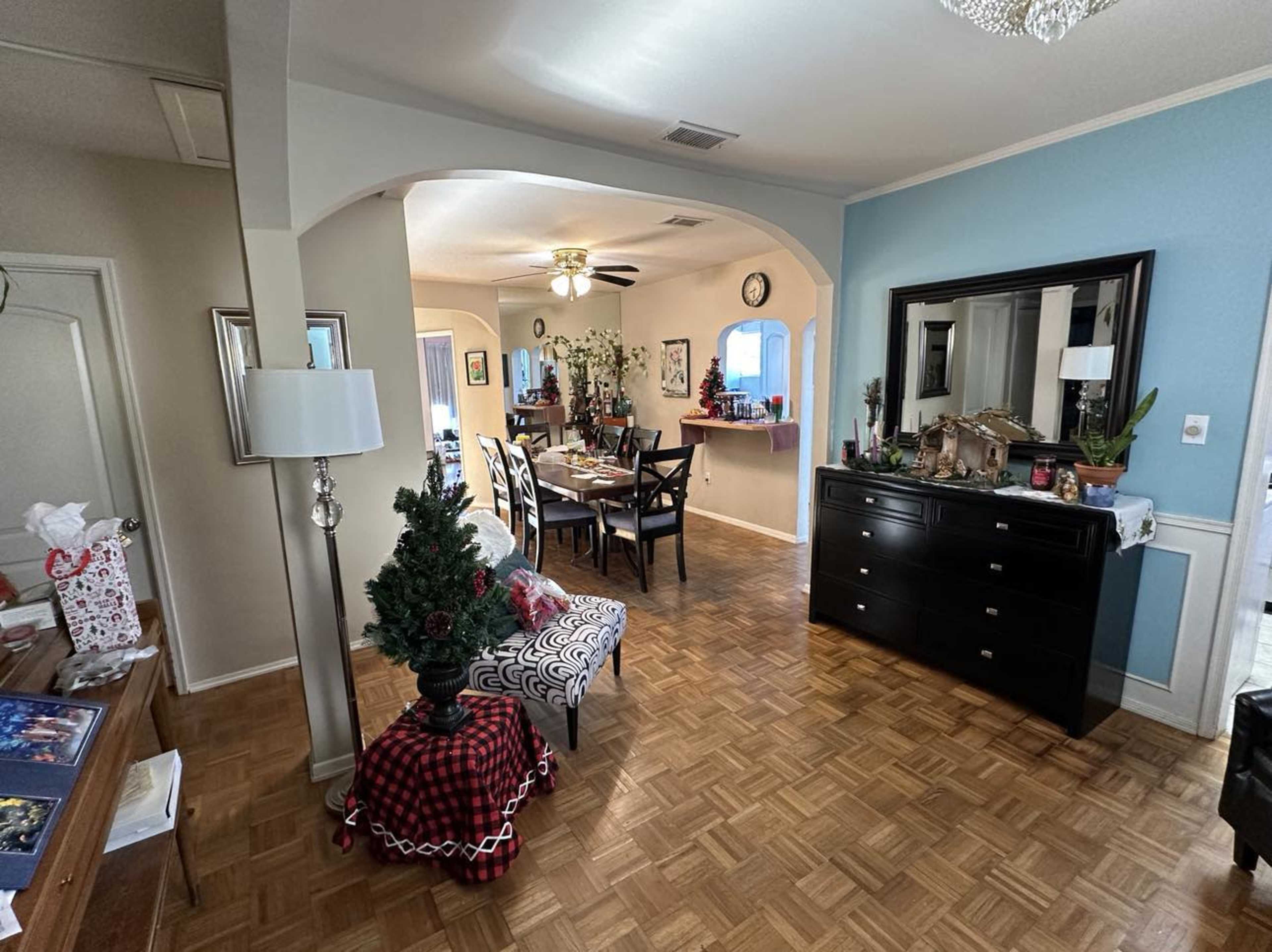 The image shows a living room with a patterned chair, a small Christmas tree, and a dining area in the background, all featuring wooden flooring.