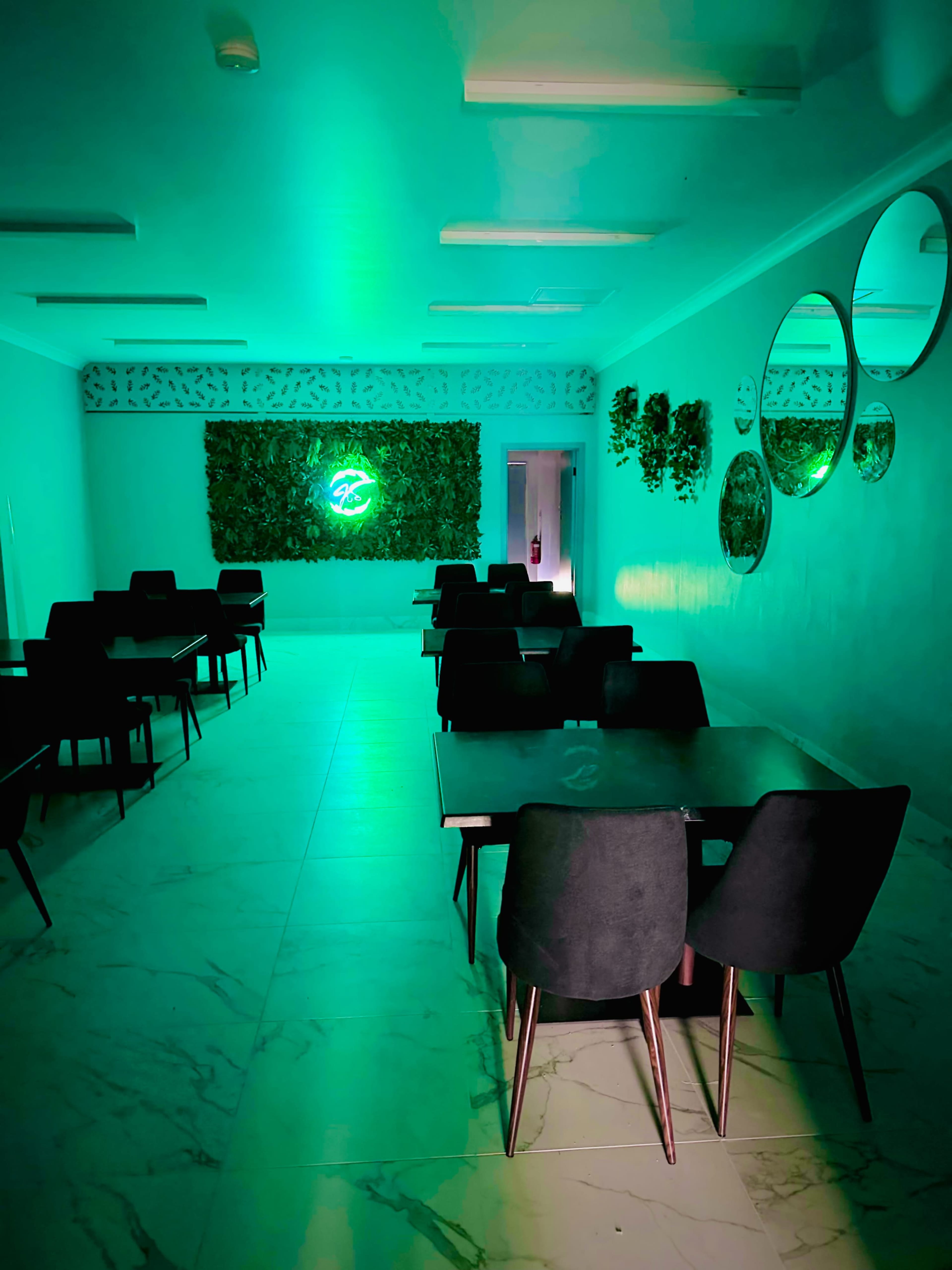 A dimly lit dining area with several black tables and chairs, decorated with green lighting and plants on one wall.
