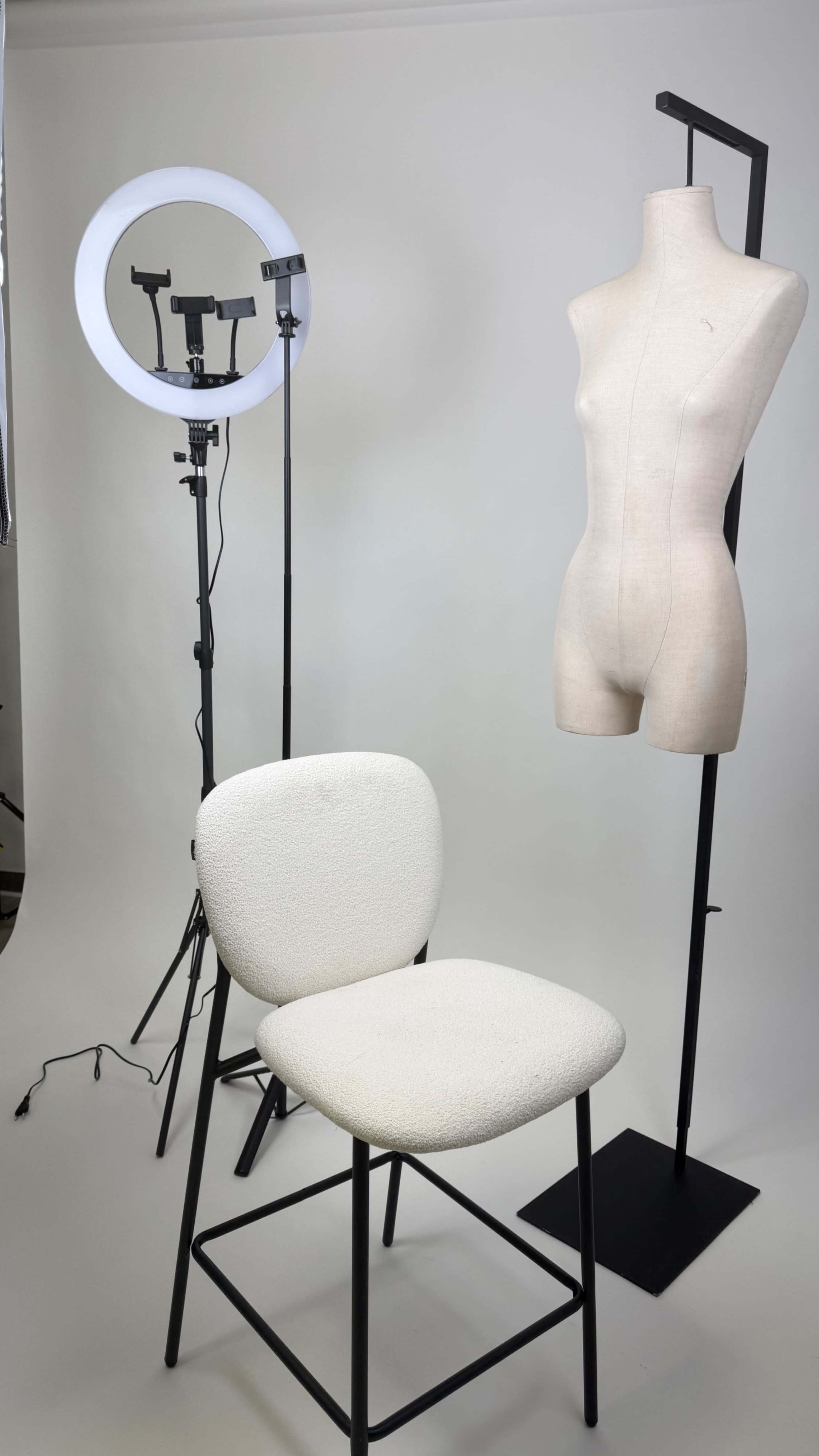 The image shows a photography setup featuring a white upholstered chair, a dress form, and a ring light on a stand, all against a neutral backdrop.
