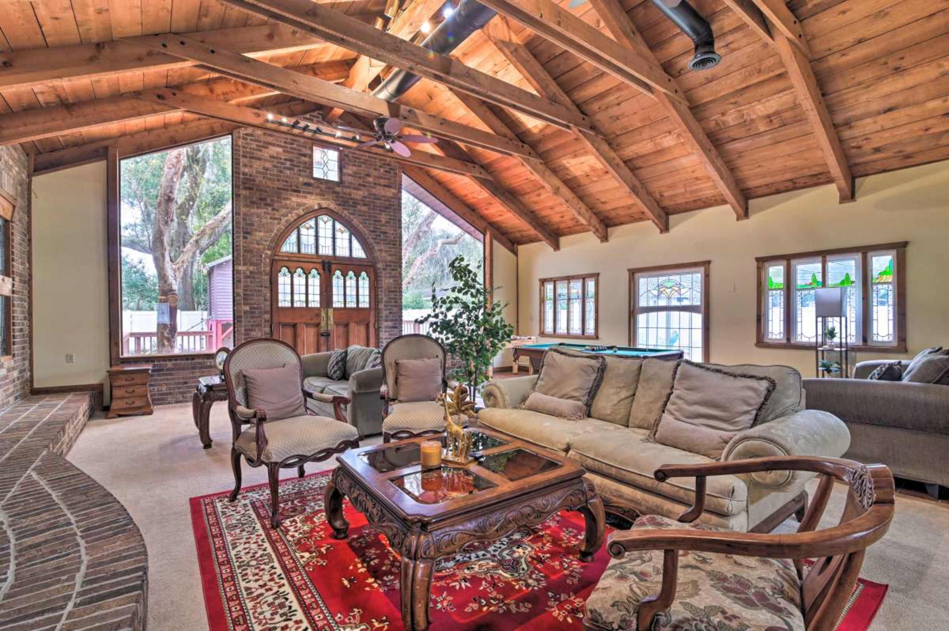 A spacious living room with a wooden ceiling, a brick fireplace, and a mix of furniture including a sofa, armchairs, and a coffee table, all arranged around a patterned rug.