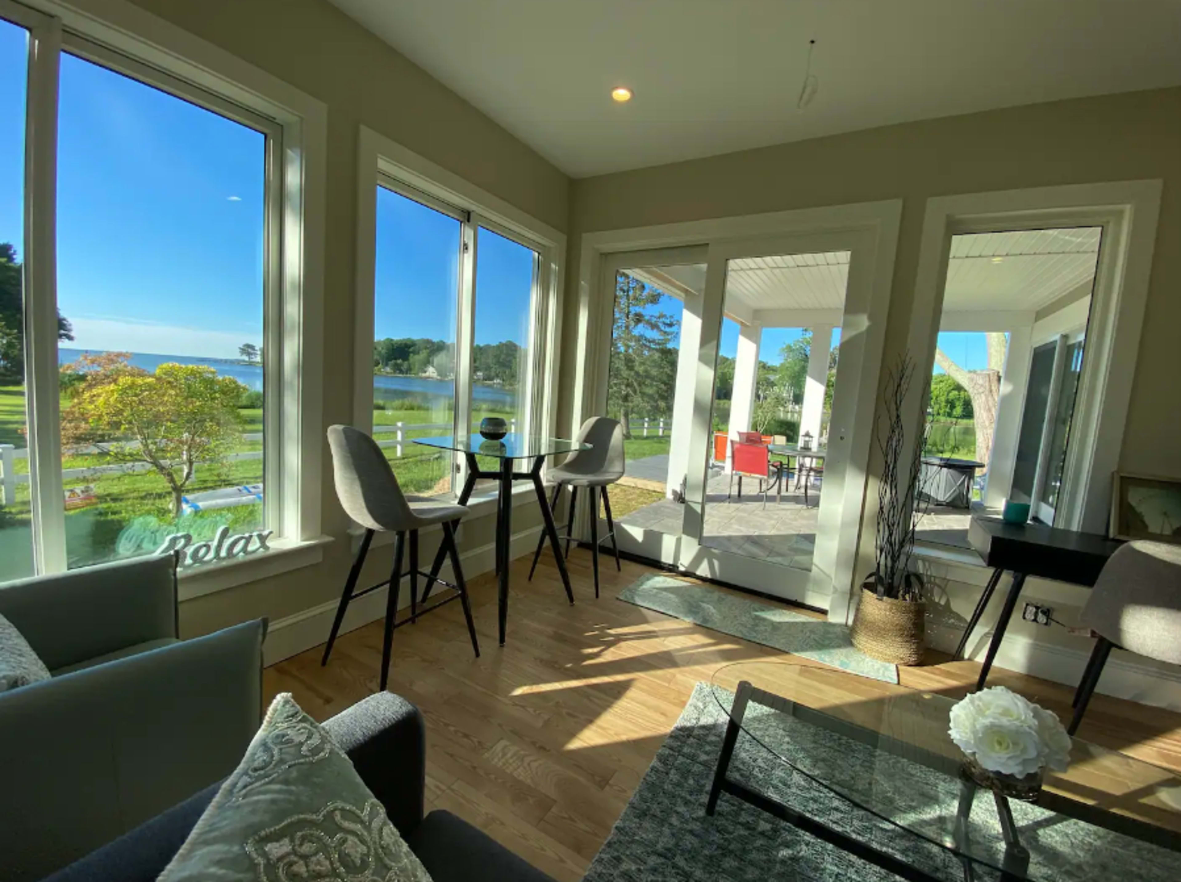 A cozy sunlit room features large windows with a view of greenery, a small dining table, and modern seating arrangements.