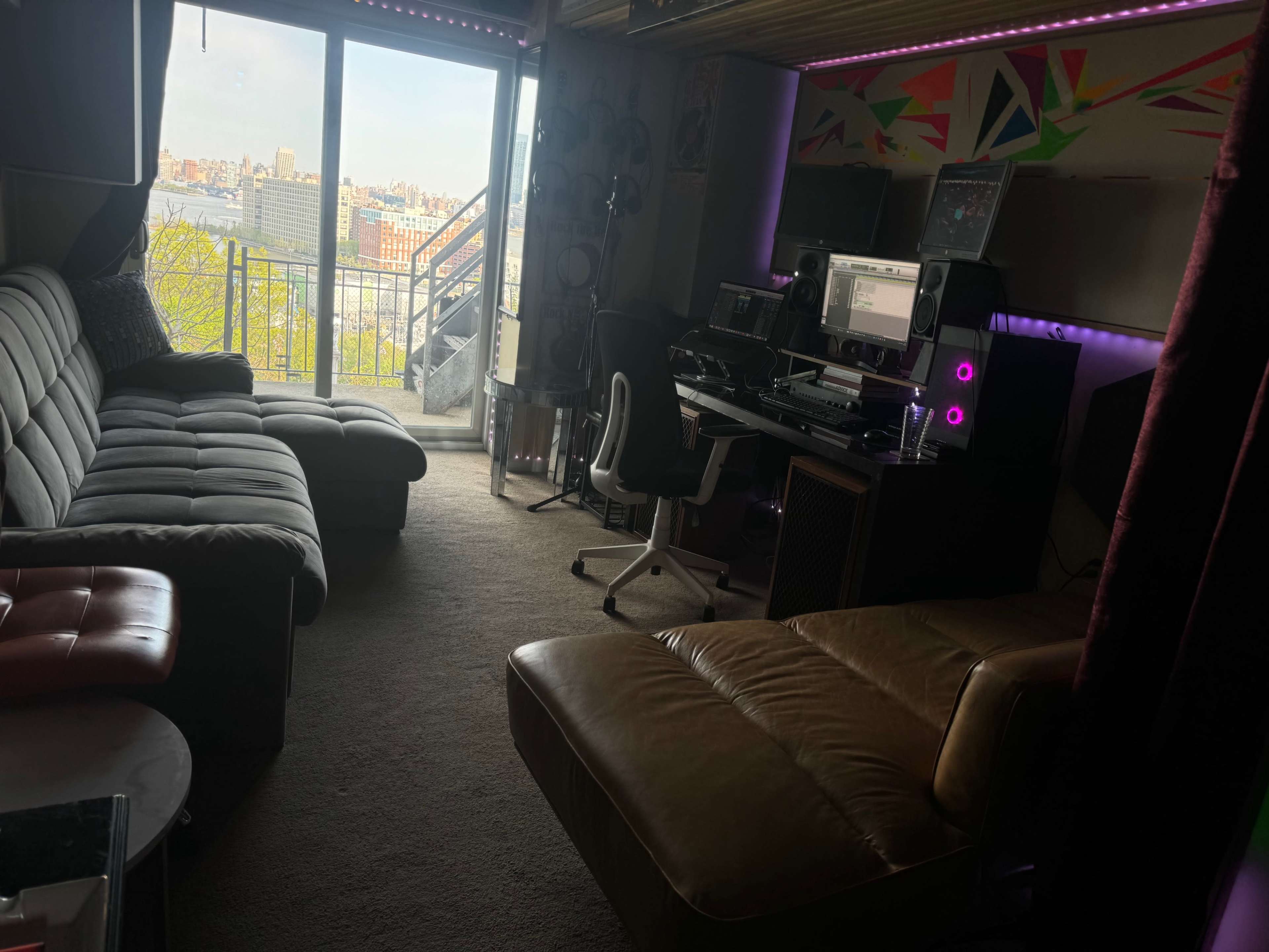 The scene shows a modern, furnished room with a cozy seating area, a desk equipped with computer equipment, and a large window revealing a cityscape outside.
