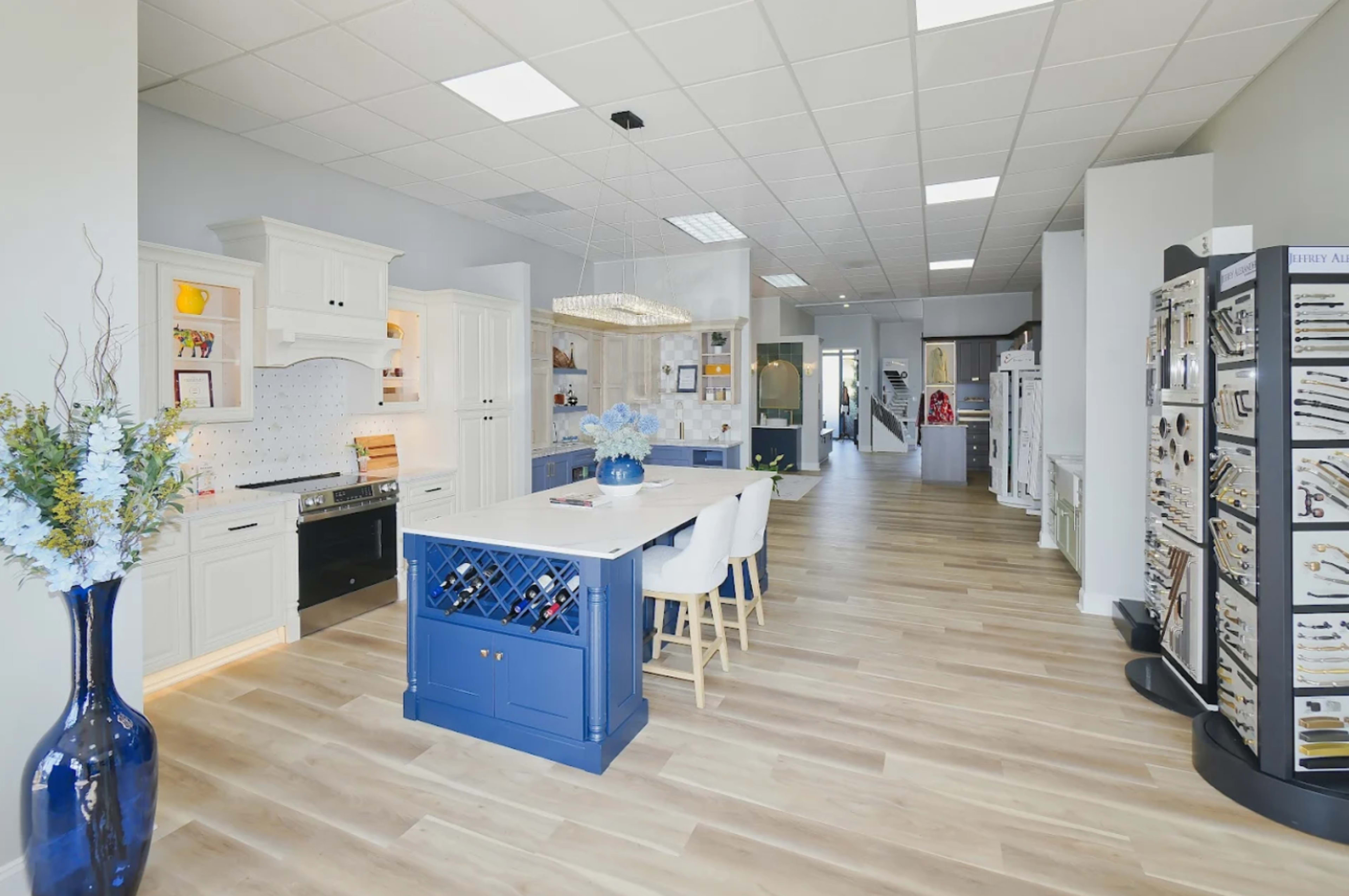 The image shows a modern kitchen showroom featuring white cabinetry, a central island with a wine rack, and a display of kitchen fixtures and hardware along the wall.