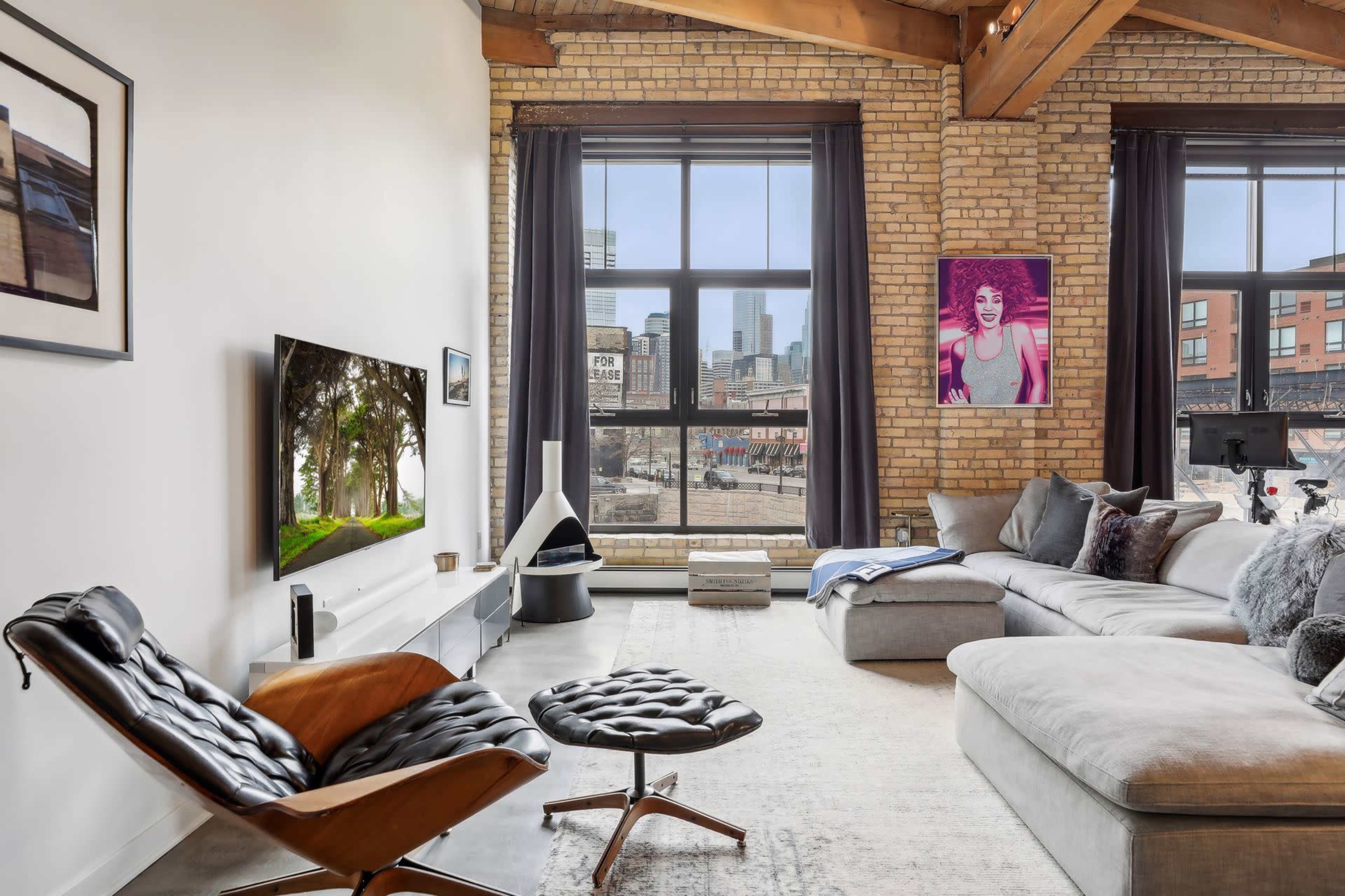 A modern living room features a sectional sofa, a sleek TV mounted on a brick wall, and large windows showcasing a city view.