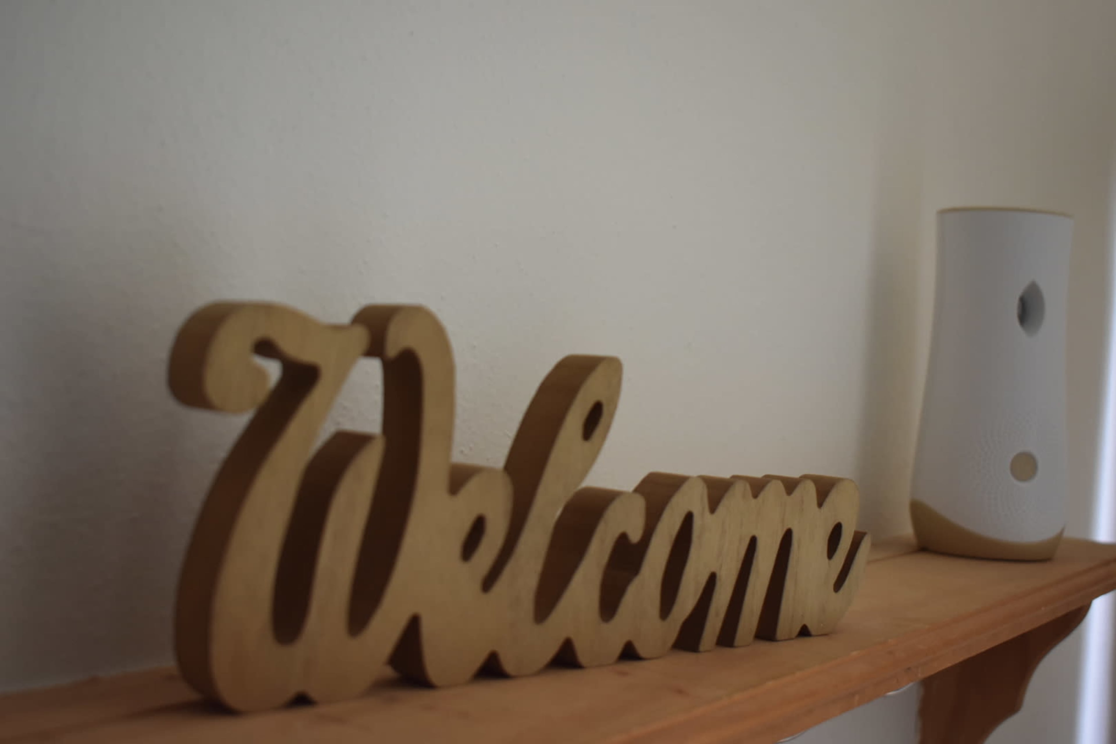 A wooden "Welcome" sign is placed on a wooden shelf beside a white decorative object.