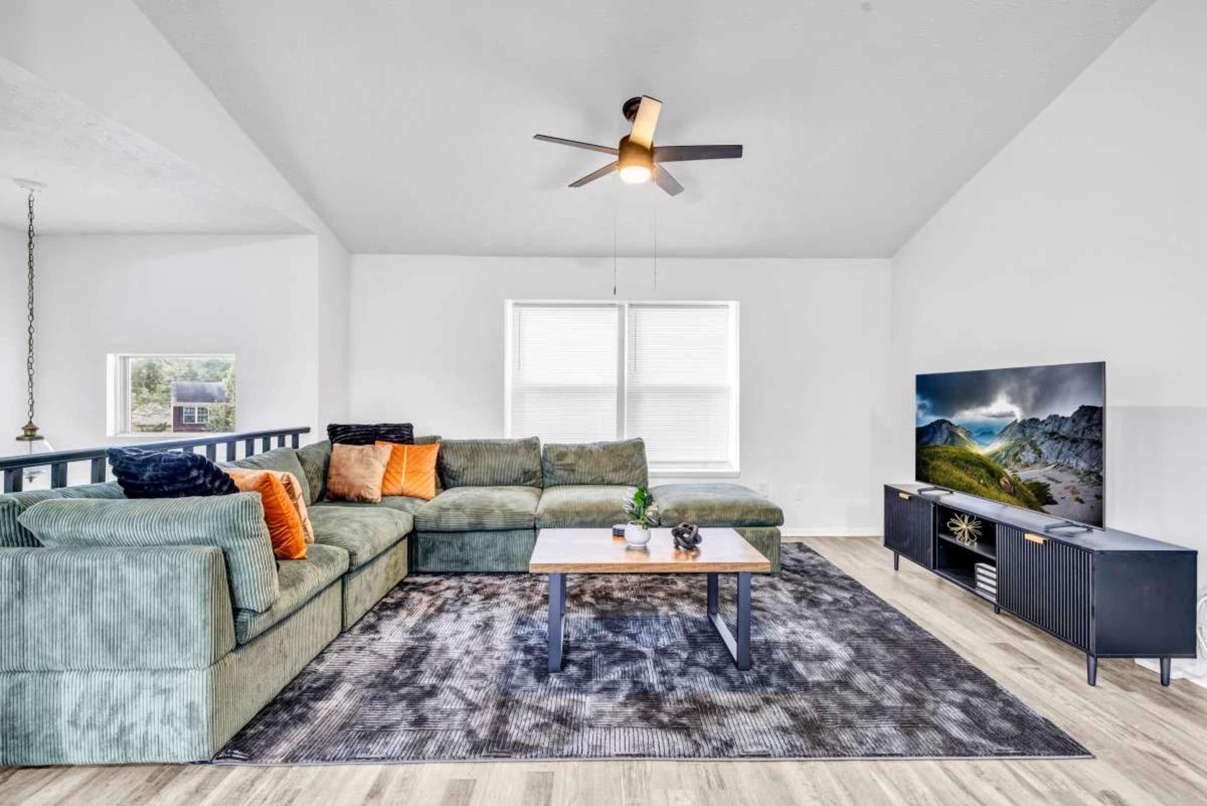 A spacious living room features a large green sectional sofa with orange cushions, a central coffee table, and a television on a black console against a light-colored wall.