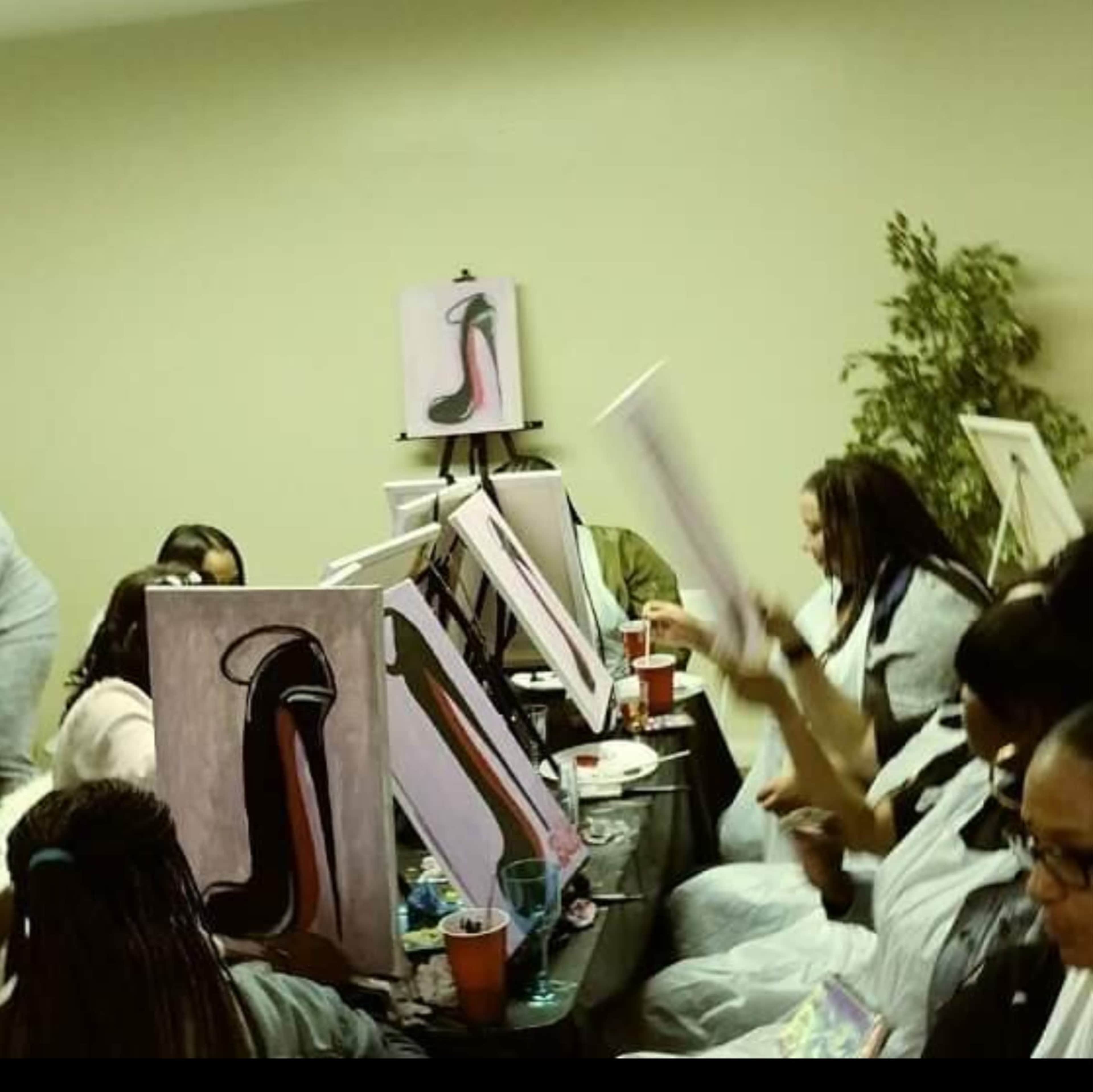 A group of people are seated at tables in a room, each working on their own painting of a high-heeled shoe.