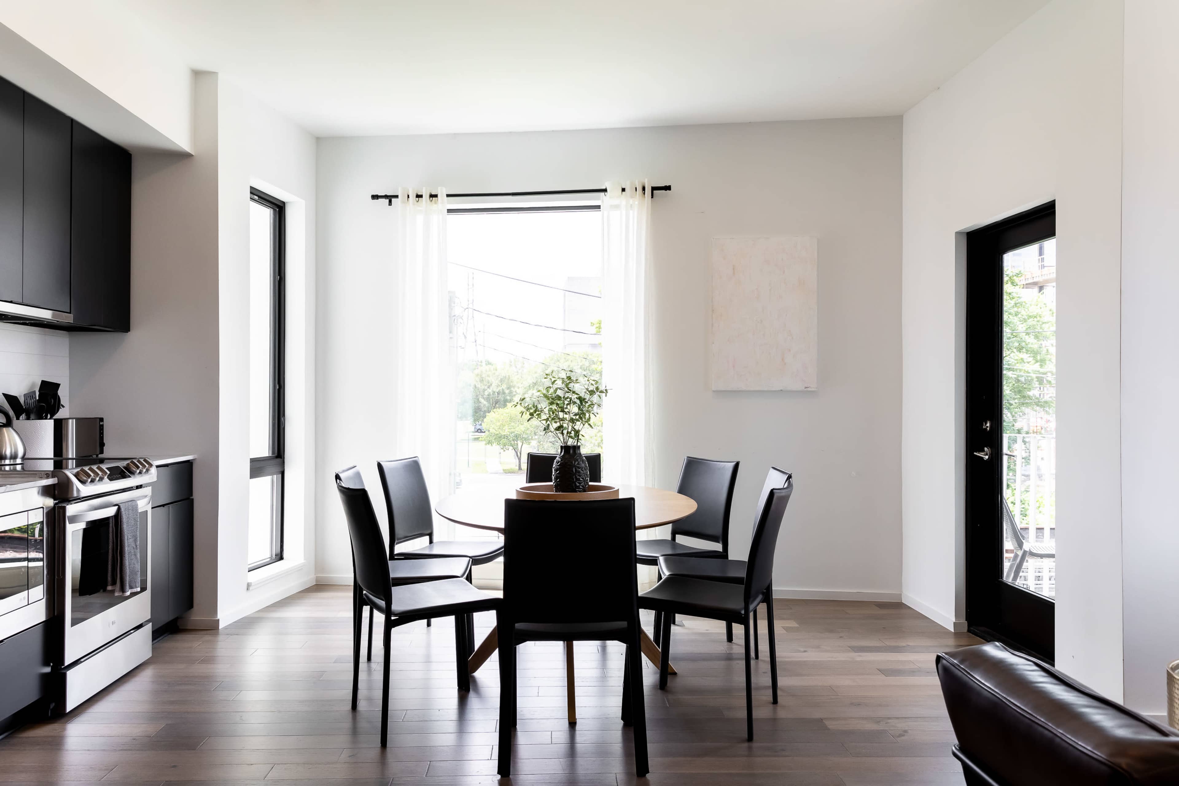 A dining area features a round table surrounded by four black chairs, with large windows letting in natural light.