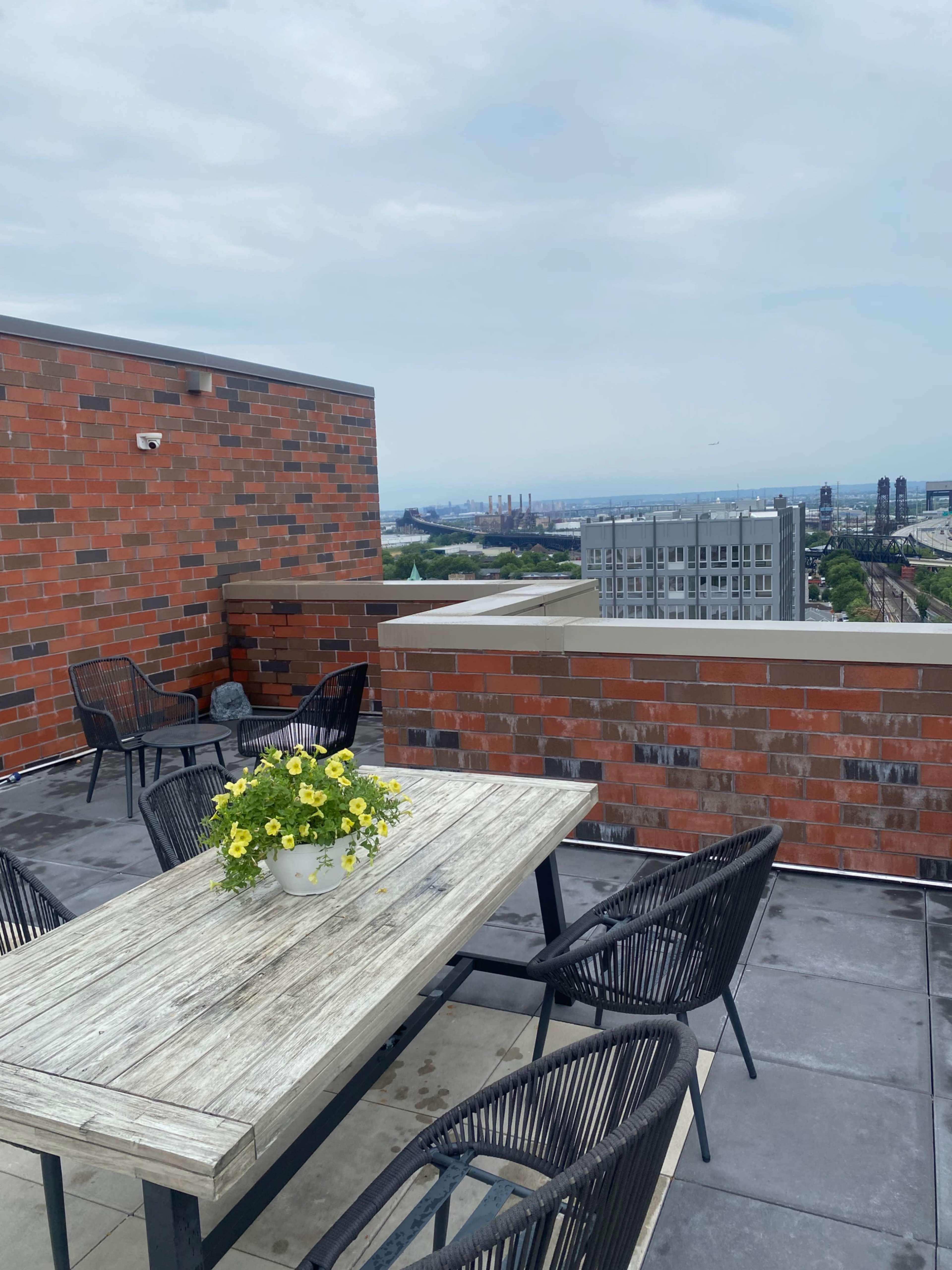 A rooftop terrace features a wooden table surrounded by black chairs and a planter of yellow flowers, overlooking a city skyline under a cloudy sky.