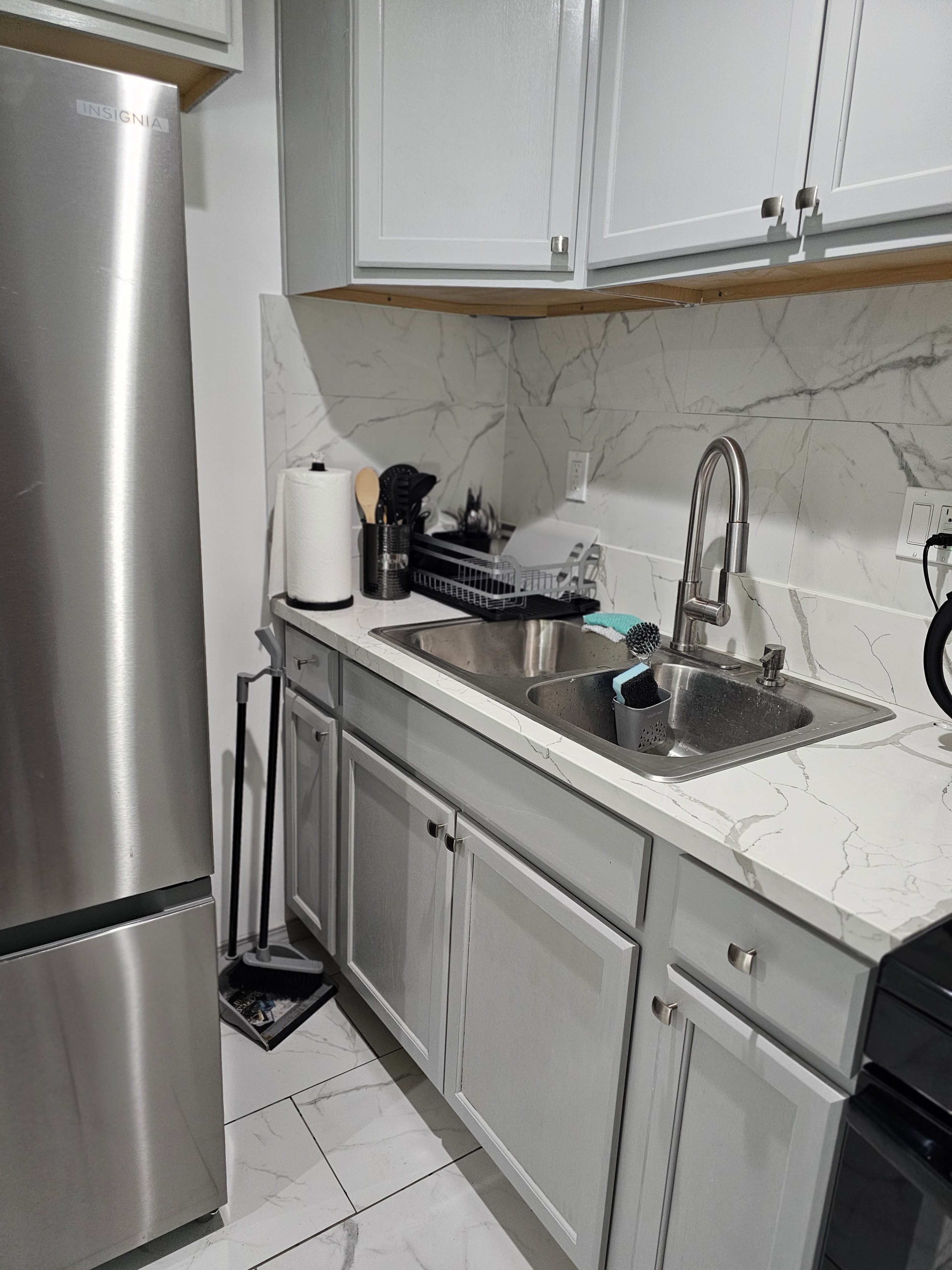 The image shows a kitchen area featuring a stainless steel refrigerator, a dual sink with a faucet, and white cabinetry, all set against a marble-patterned backsplash.