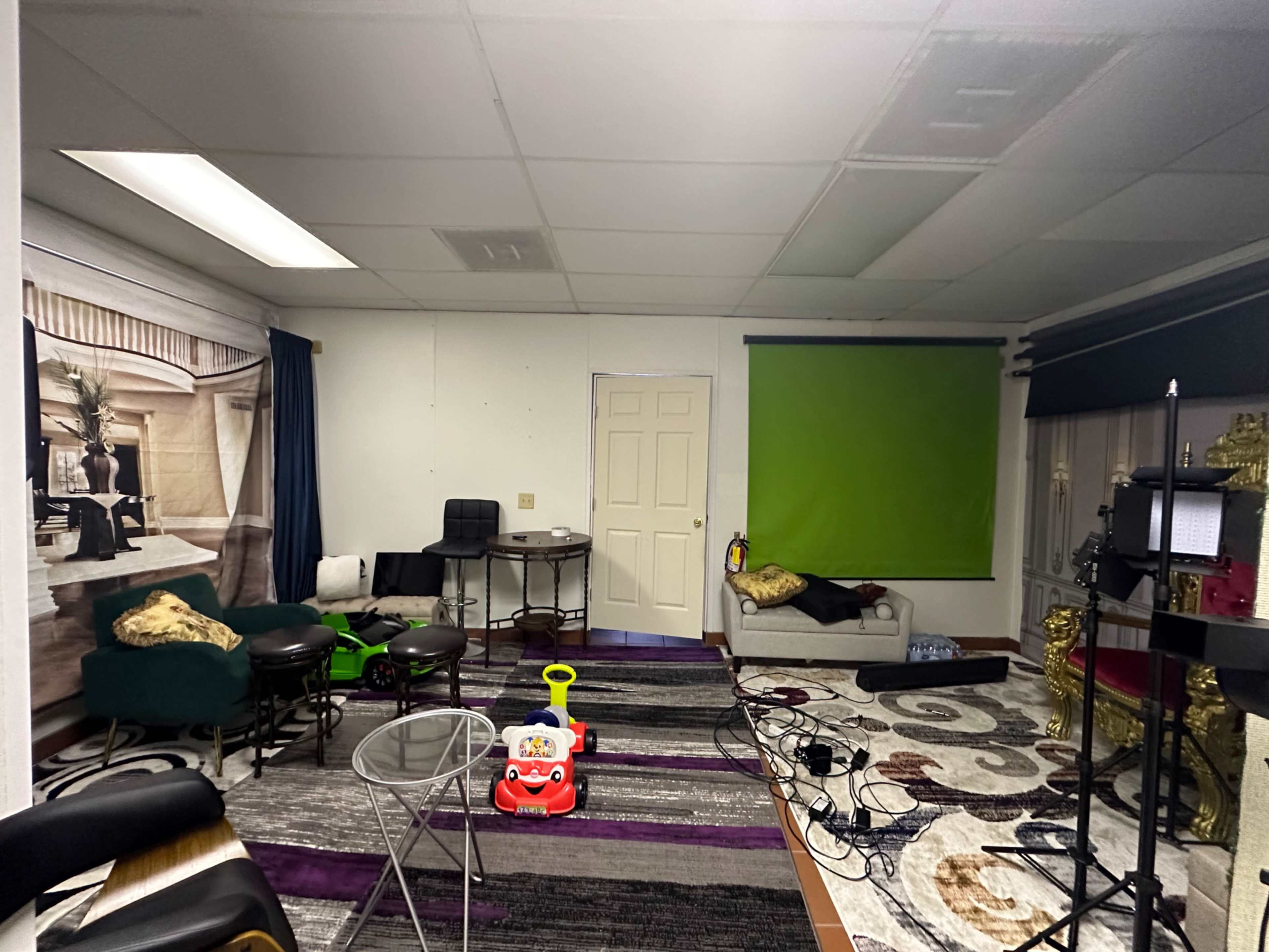 The image shows a partially furnished room with a green screen, various chairs, a toy ride-on car, and a patterned rug.