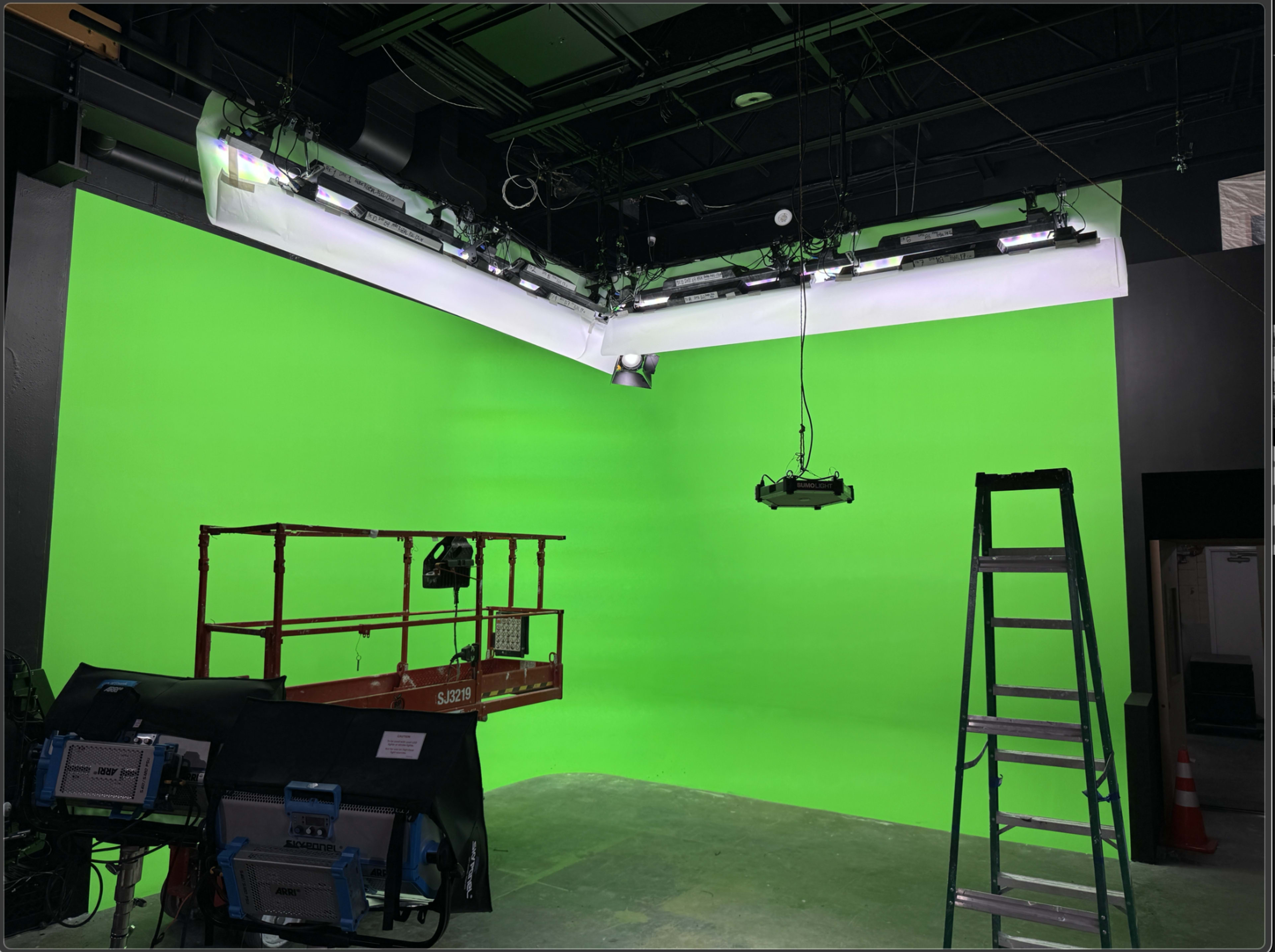 The image shows a studio with a green screen backdrop, a ladder, and a scissor lift positioned in front of it.