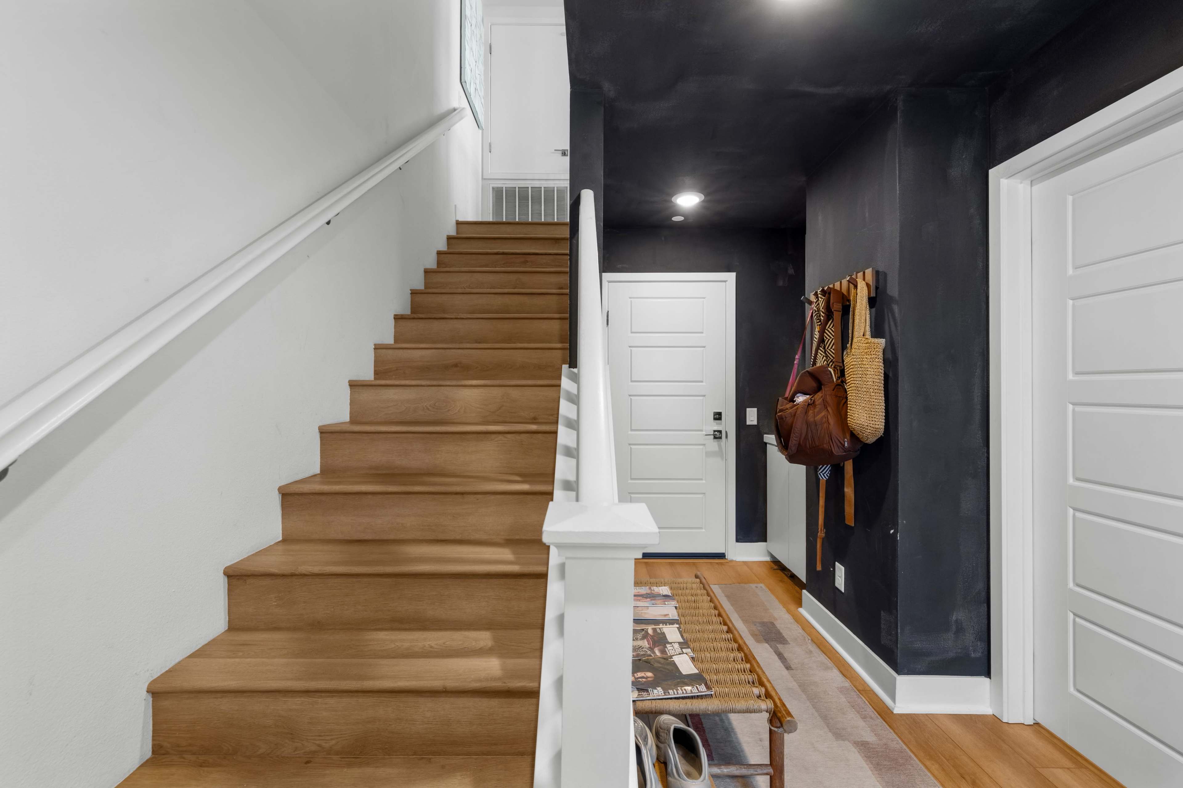 The image shows a well-lit hallway leading to a staircase with wooden steps, white walls, and a dark accent wall, featuring a bench and bags hanging on hooks.