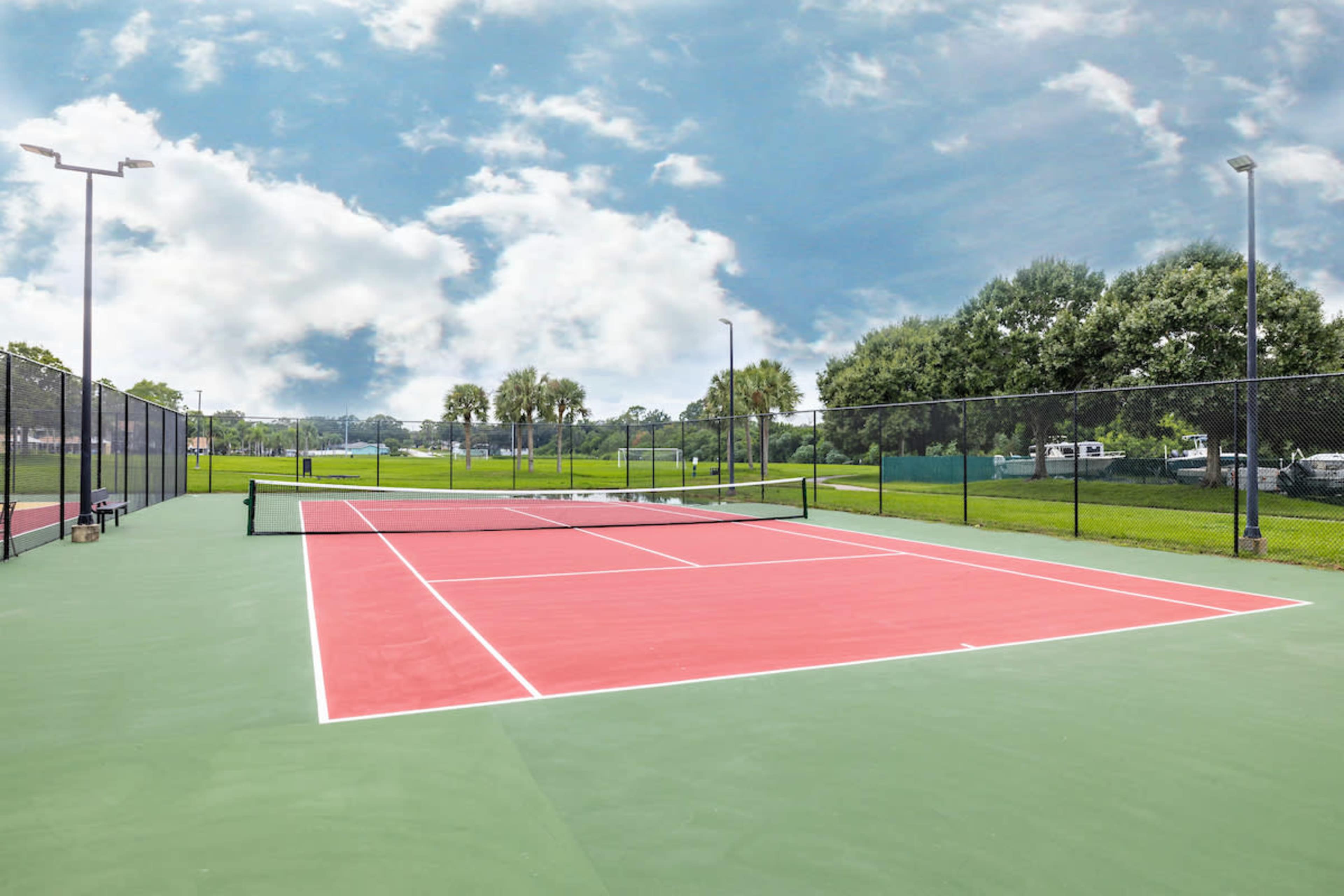 A well-maintained tennis court with red and green surfaces under a partly cloudy sky.