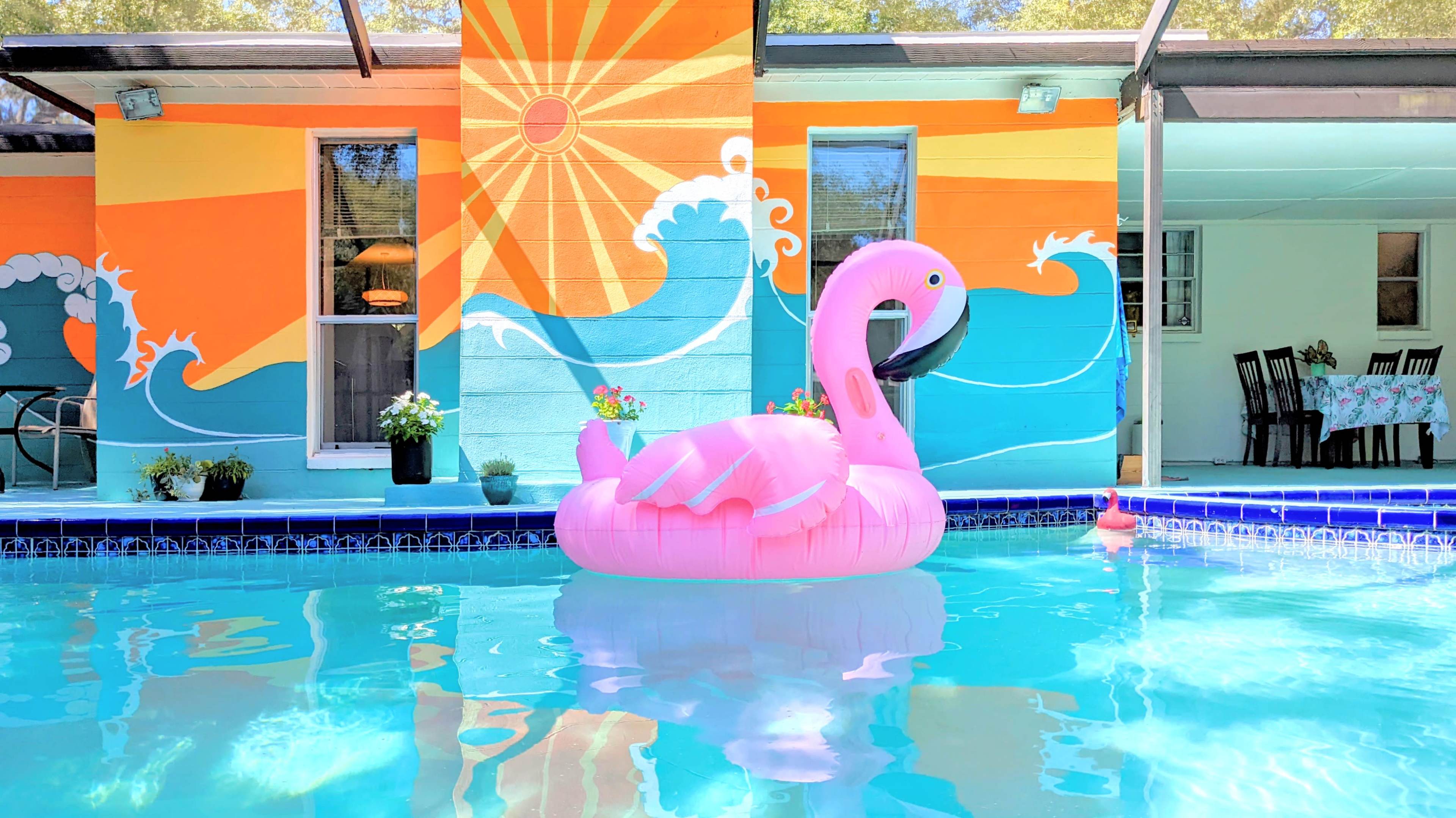 A large pink flamingo float sits in a blue swimming pool with a colorful mural of waves and a sun on the wall behind it.