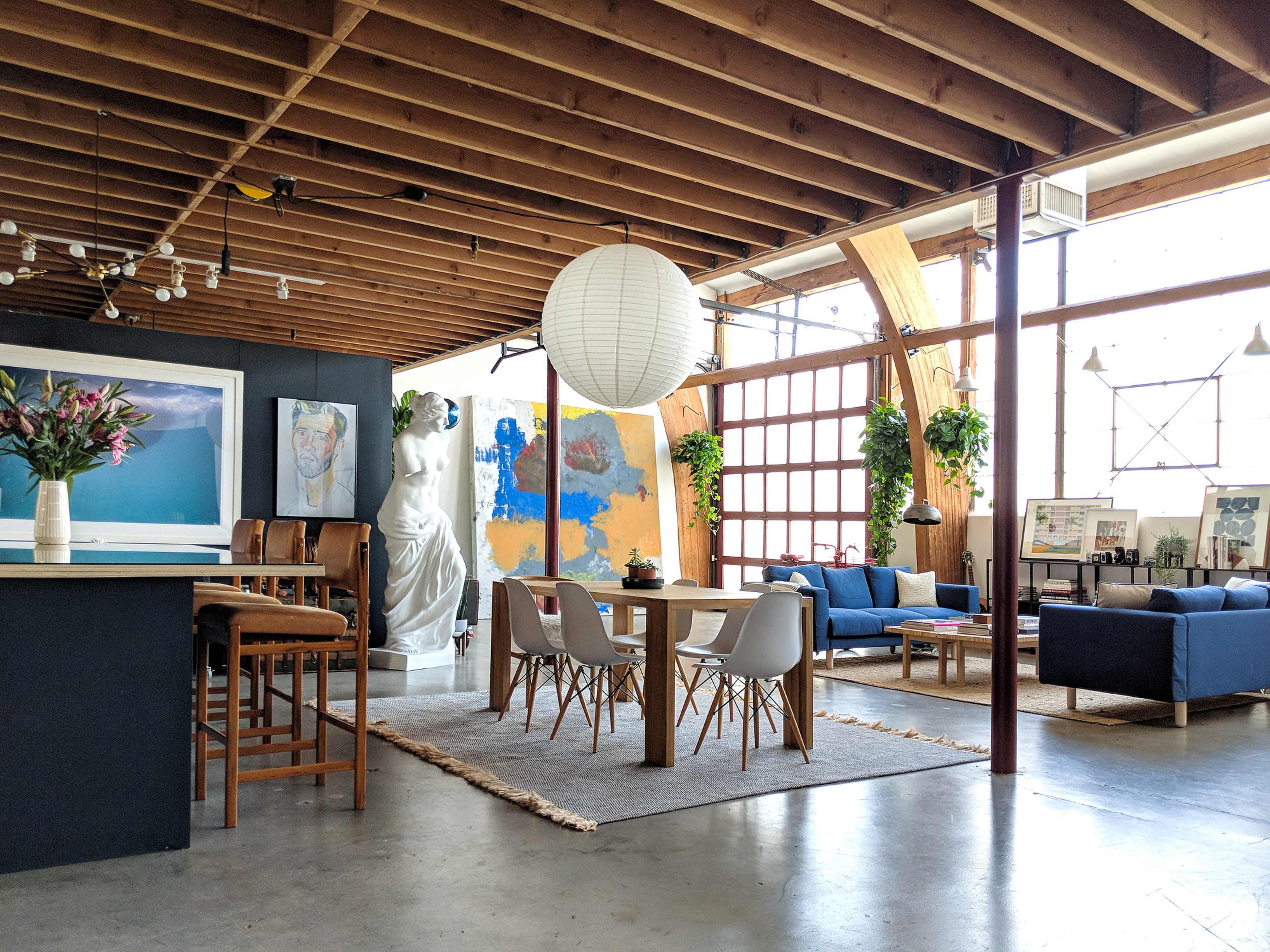 A stylish open-concept living space featuring a dining area with a large table, a kitchen bar, a sculpture, and various plants around the room.