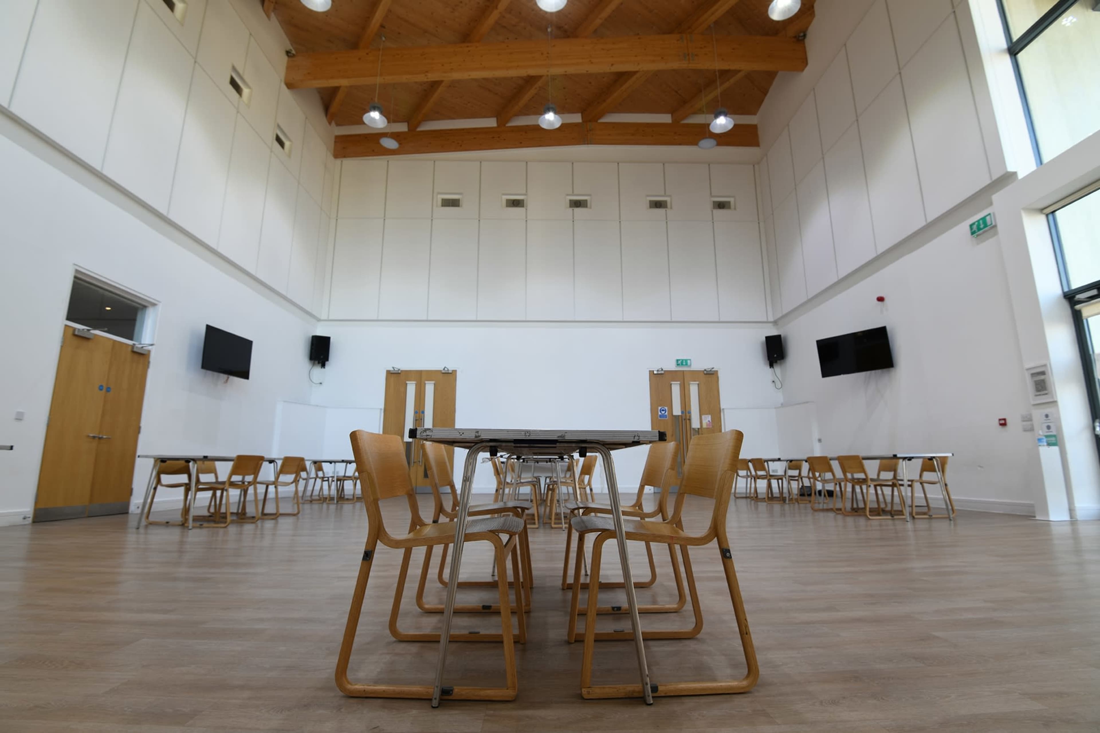 A spacious, empty room with wooden ceiling beams, multiple tables with chairs stacked on them, and television screens mounted on the walls.