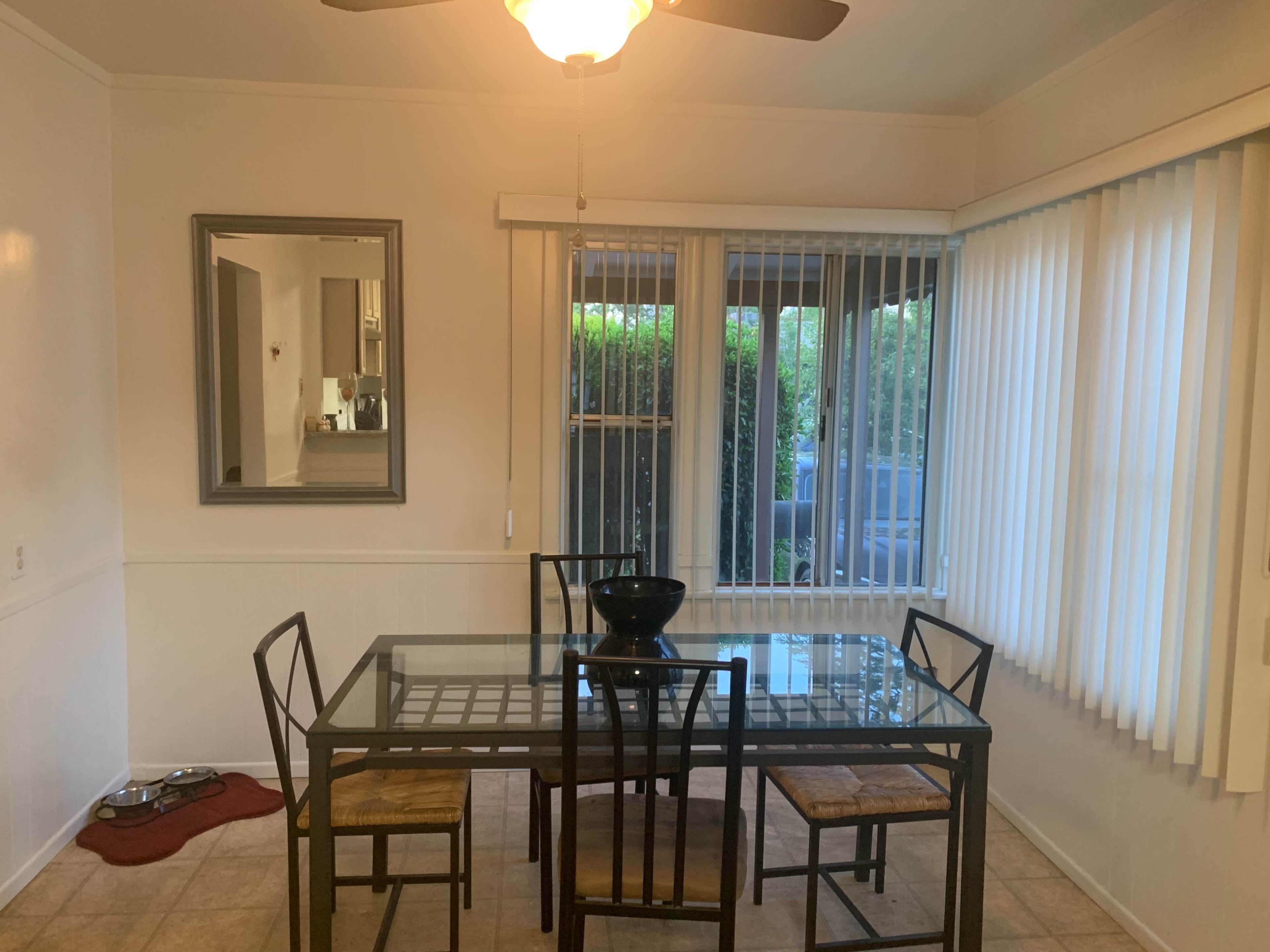 A glass-top dining table and chairs are set in a well-lit, neutral-toned dining area with large windows and vertical blinds.