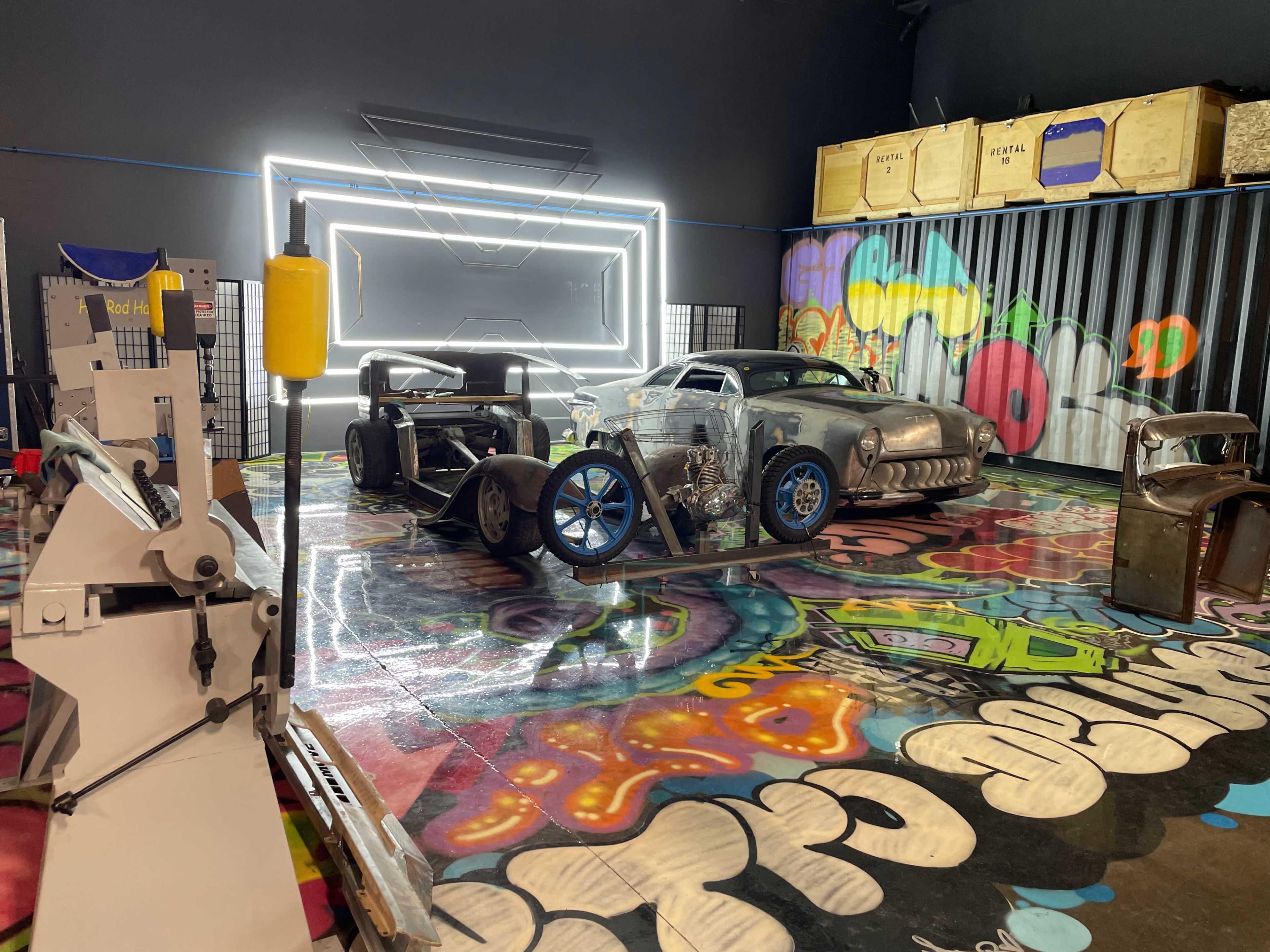 The image shows two custom cars situated on a colorful, graffiti-covered floor in a garage setting, with neon lighting in the background.