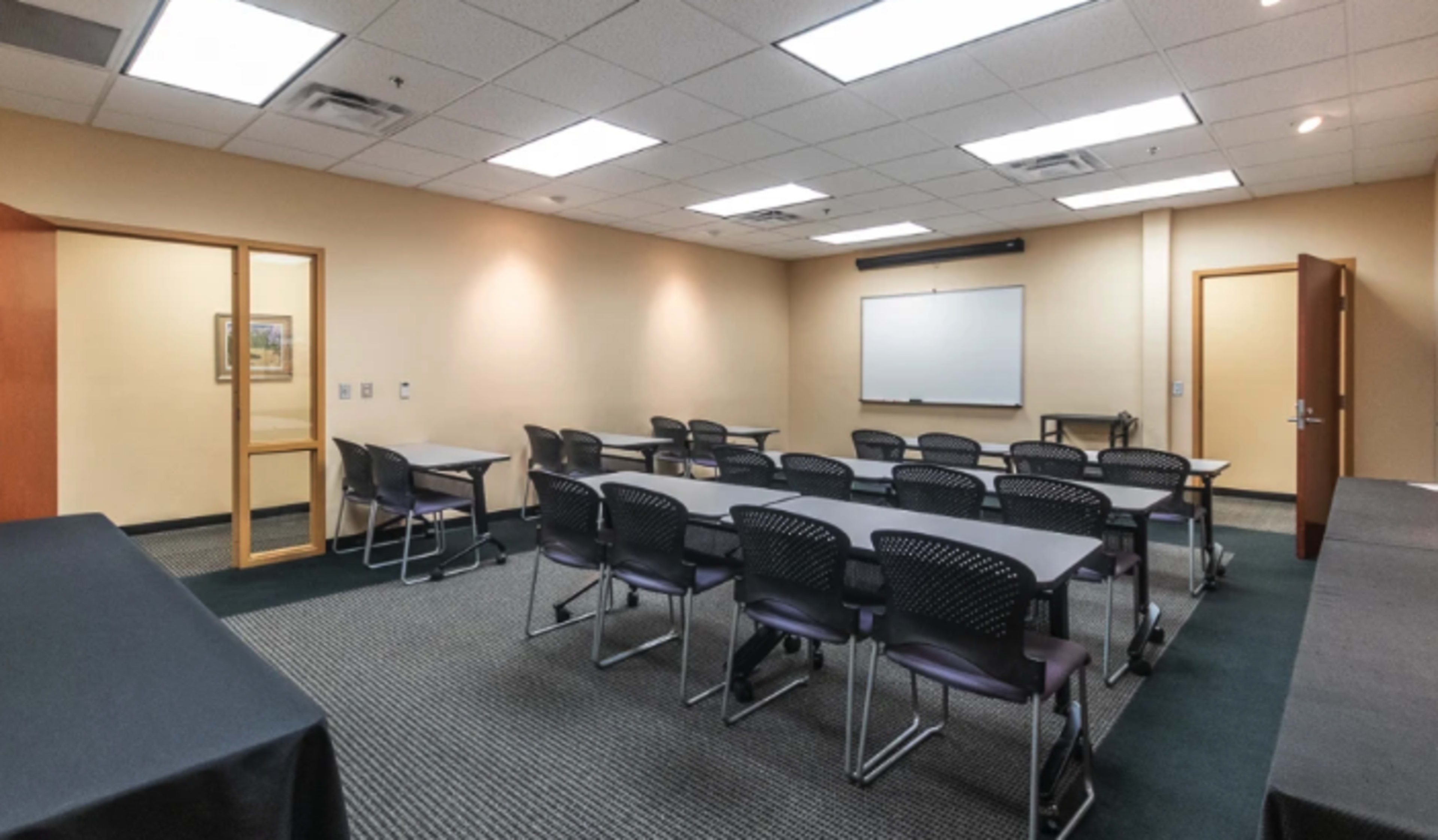 A meeting room arranged with several tables and chairs, featuring a whiteboard and a door leading to another space.