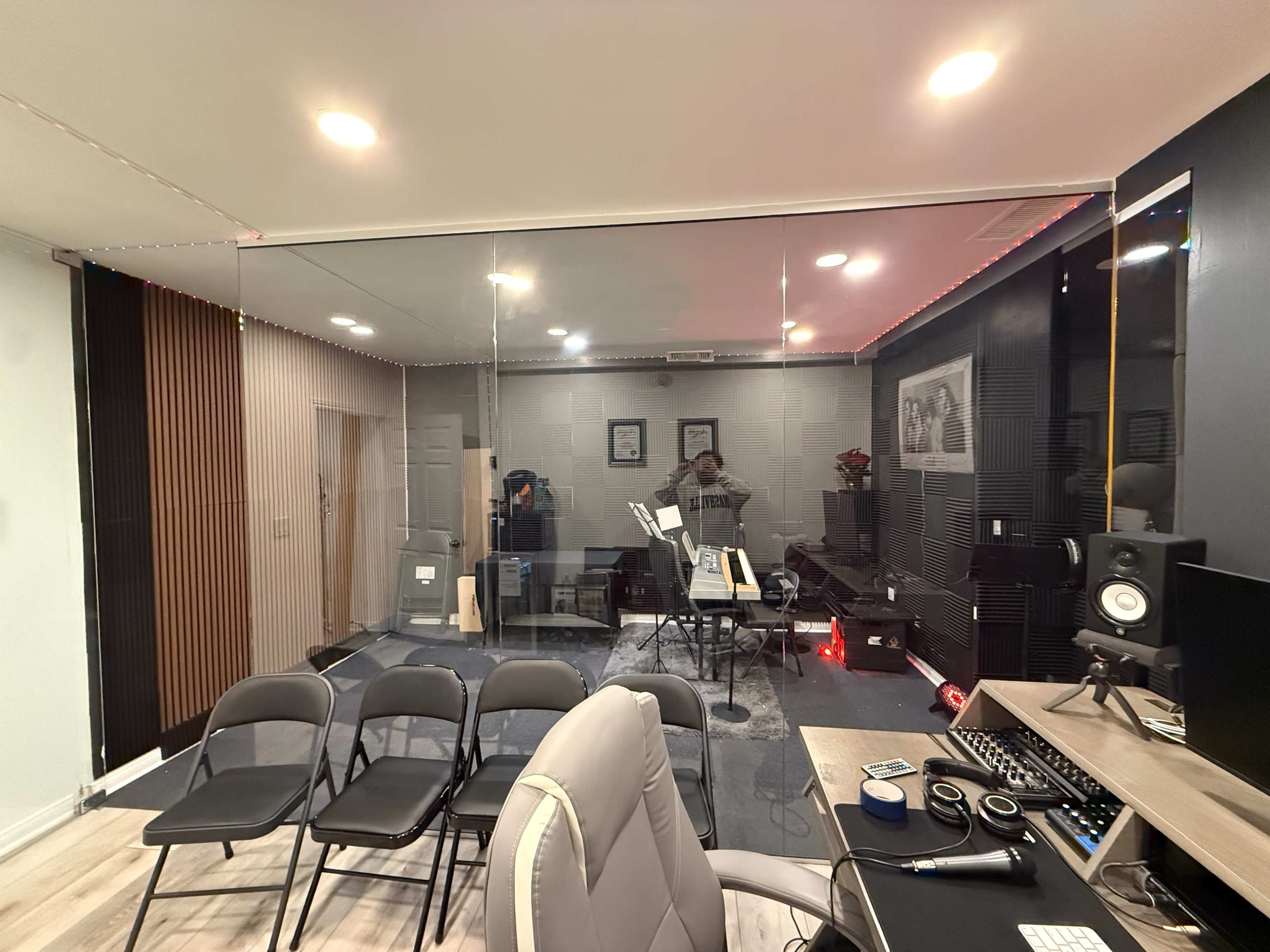 The image shows a music studio with a glass wall, featuring instruments and equipment inside, and several empty chairs in the foreground.