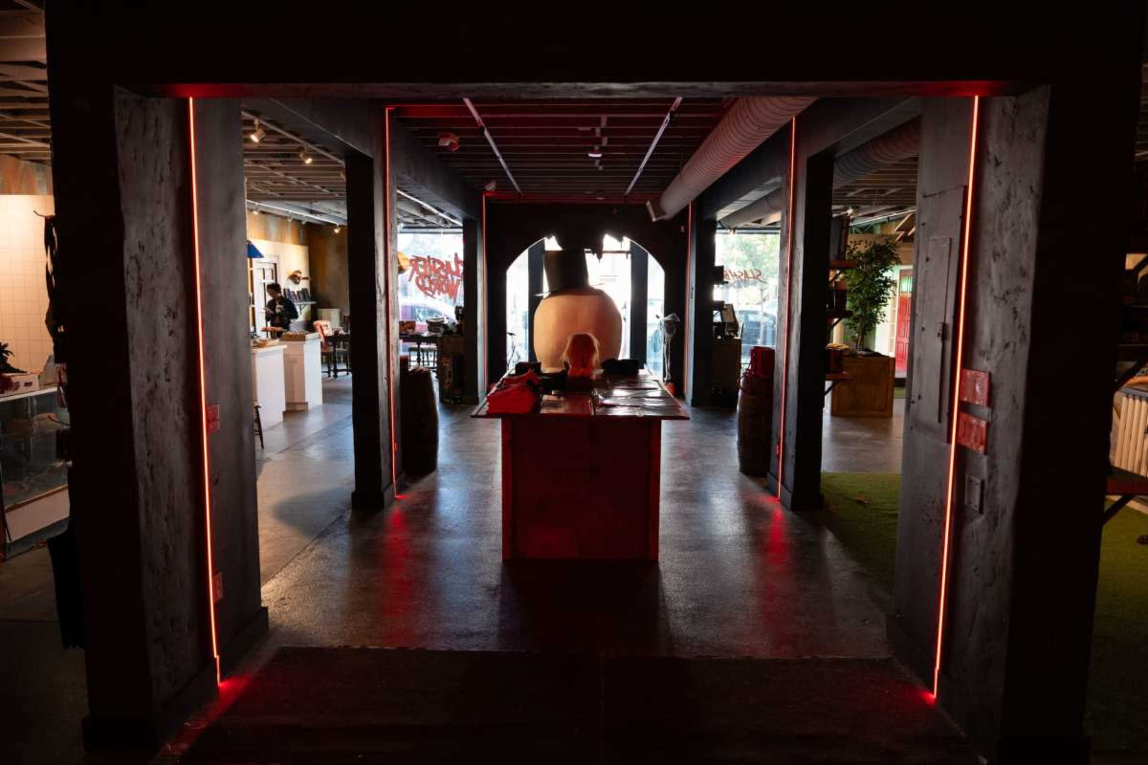 The image shows a dimly lit space with a central counter under soft red lights, flanked by dark columns and a large white sculpture in the background.