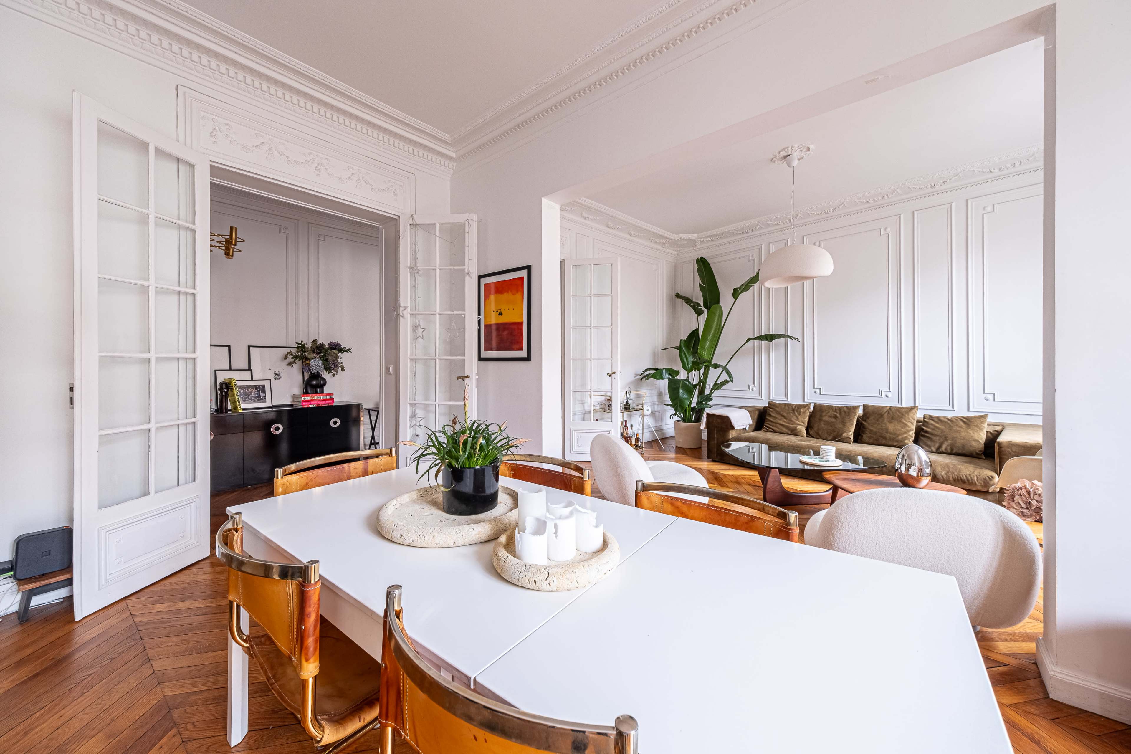 A spacious dining area features a white table surrounded by clear and beige chairs, with an adjacent living room displaying a green couch and large houseplants.