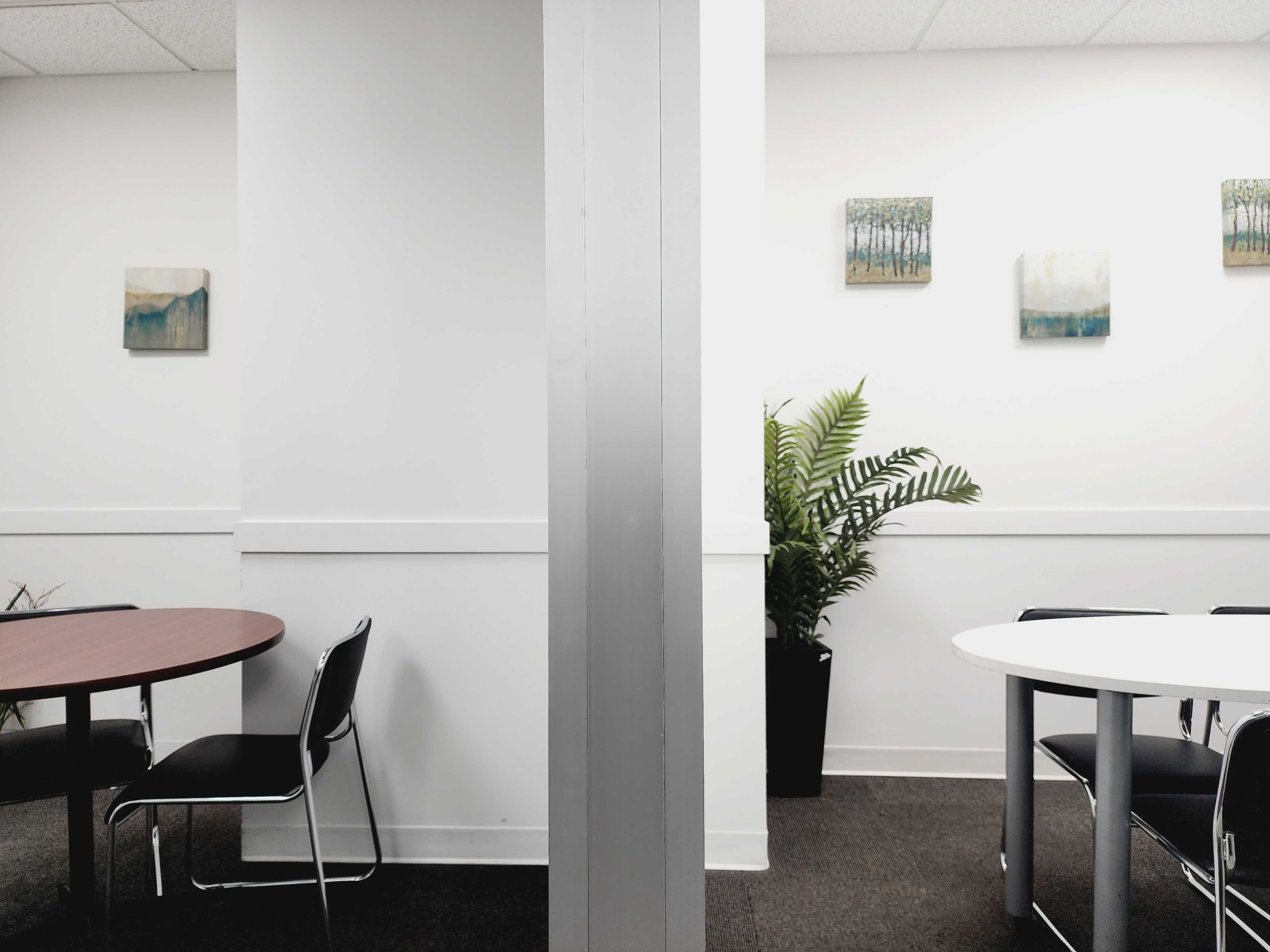 The image shows a split view of an office space with two tables and a potted plant against a white wall adorned with artworks.