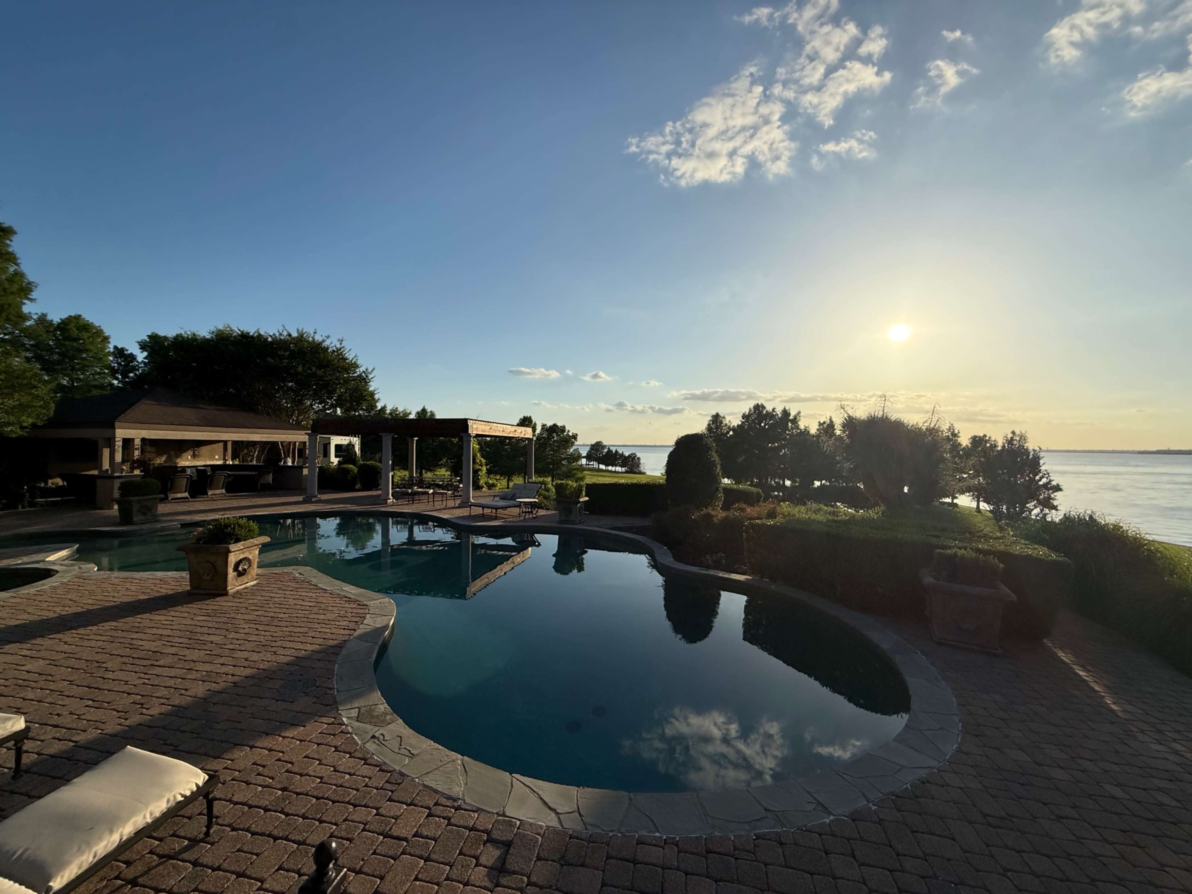 The image shows a scenic backyard with a swimming pool, surrounded by green trees and a sunset over a body of water.