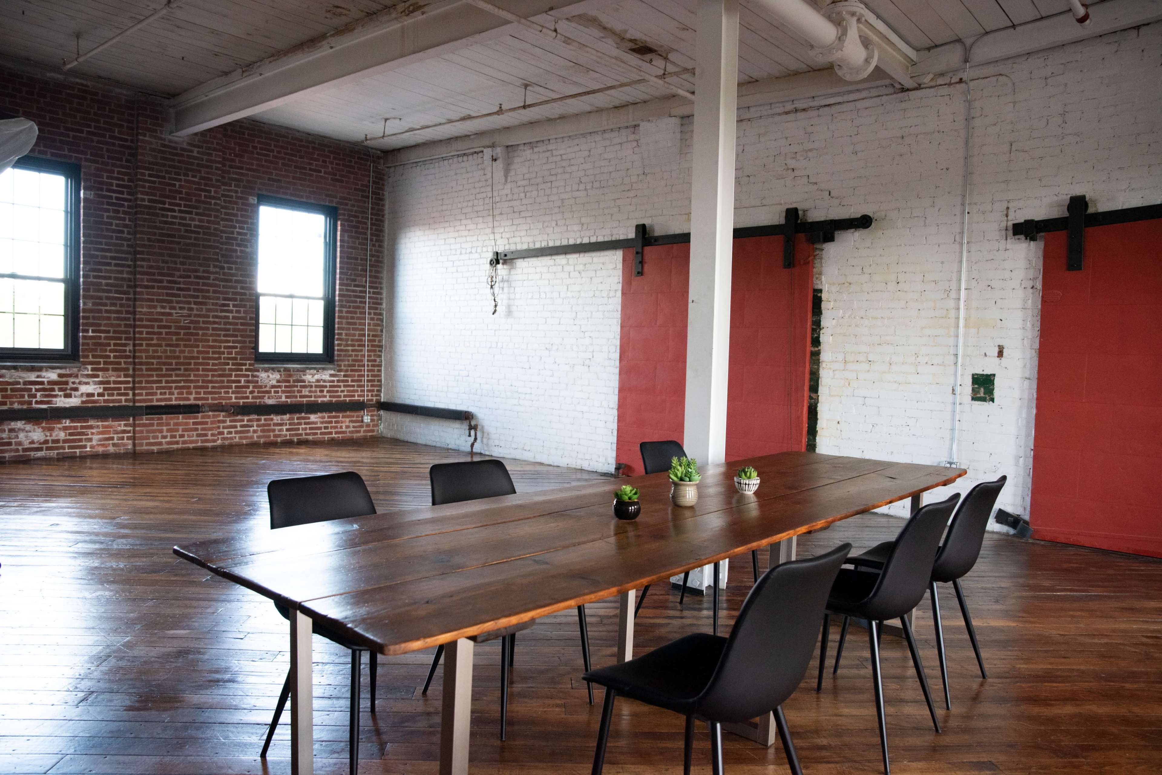 A large wooden table with chairs surrounds it in an industrial-style room featuring brick walls and red sliding doors.
