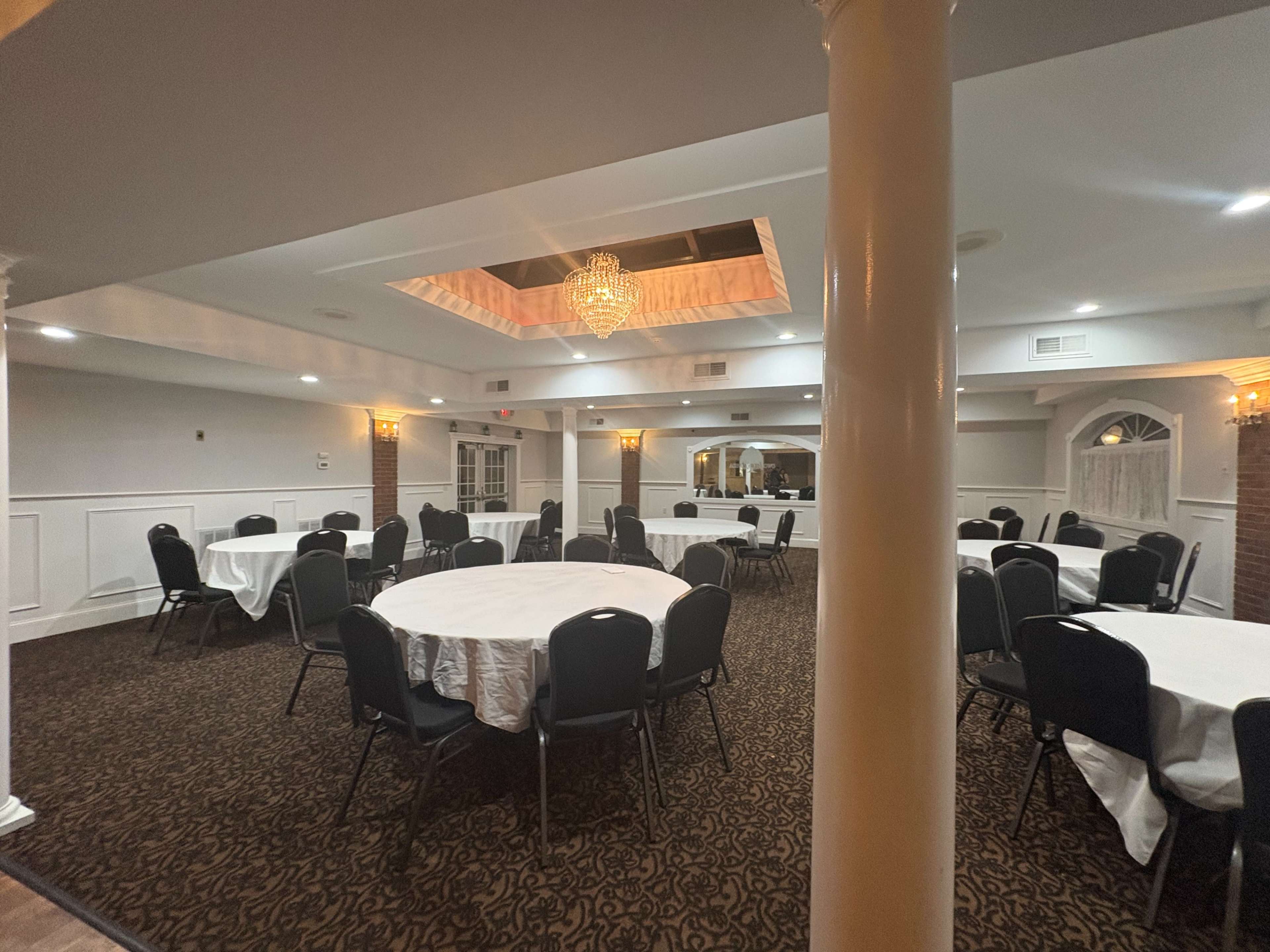 The image shows a banquet room with several round tables covered in white tablecloths, surrounded by black chairs, and featuring a chandelier overhead.